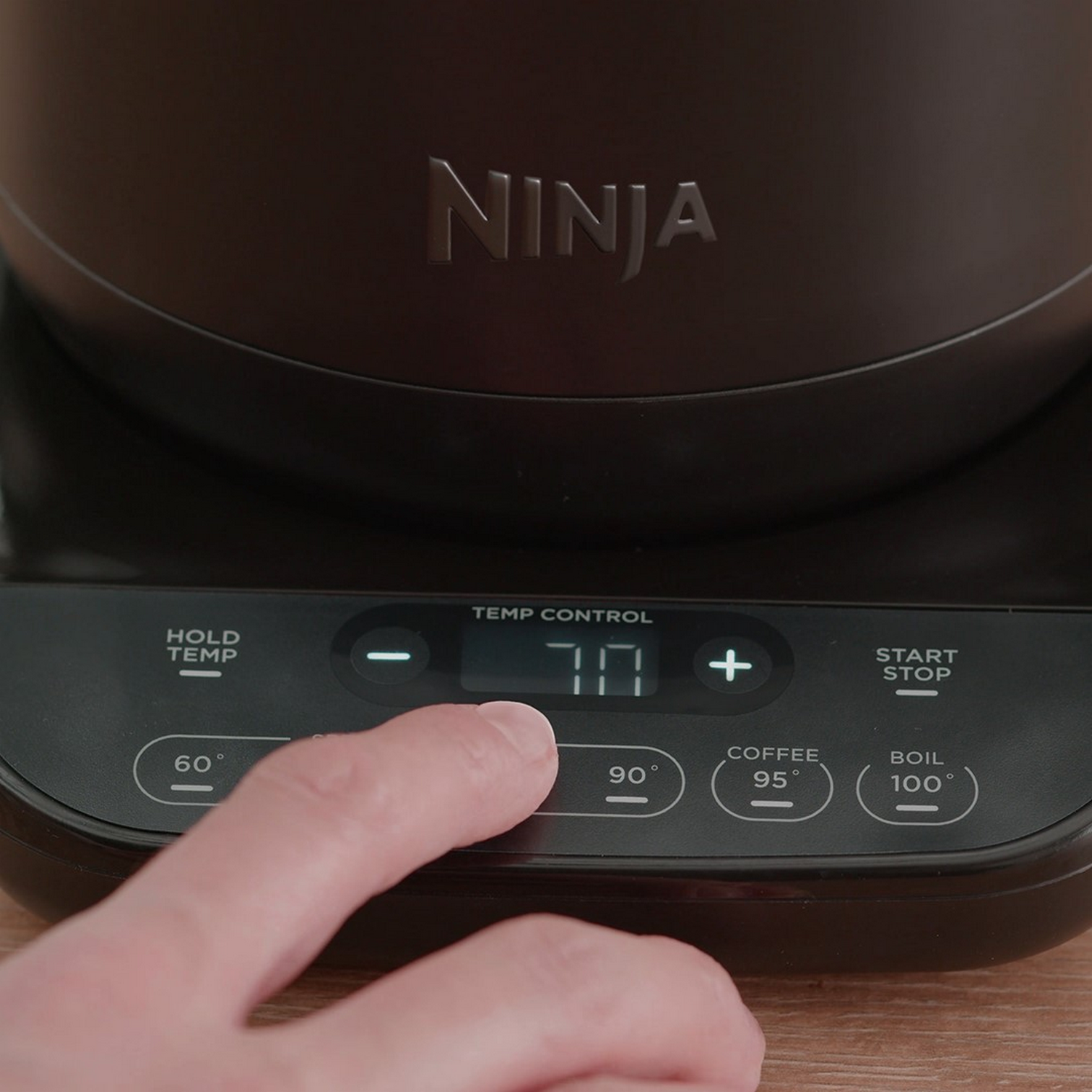 Ręka naciska przycisk ekspresu do kawy Ninja, wyświetlający 70 stopni. Czarna podstawa, wyświetlacz cyfrowy i przyciski sterowania.