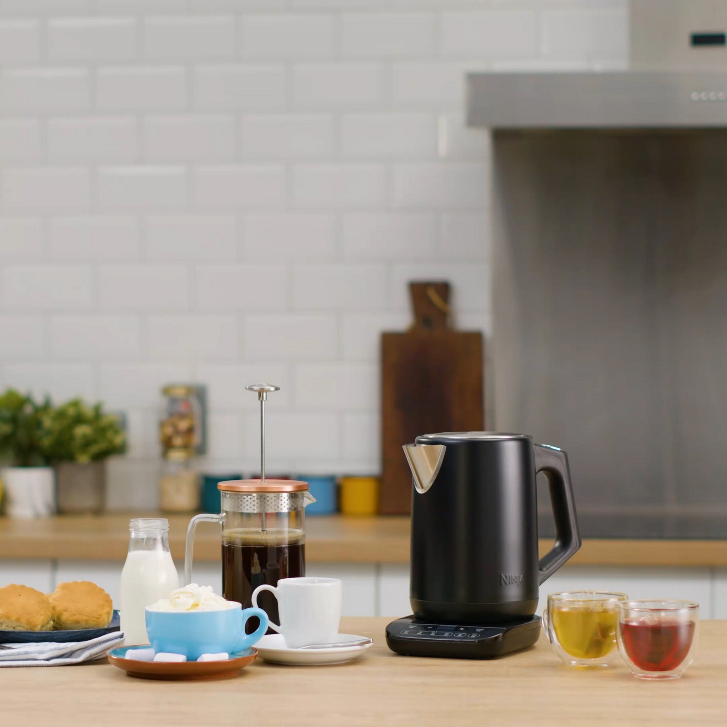 Scena kuchenna z ekspresem do kawy, czajnikiem, filiżankami i ciastami na drewnianym stole.