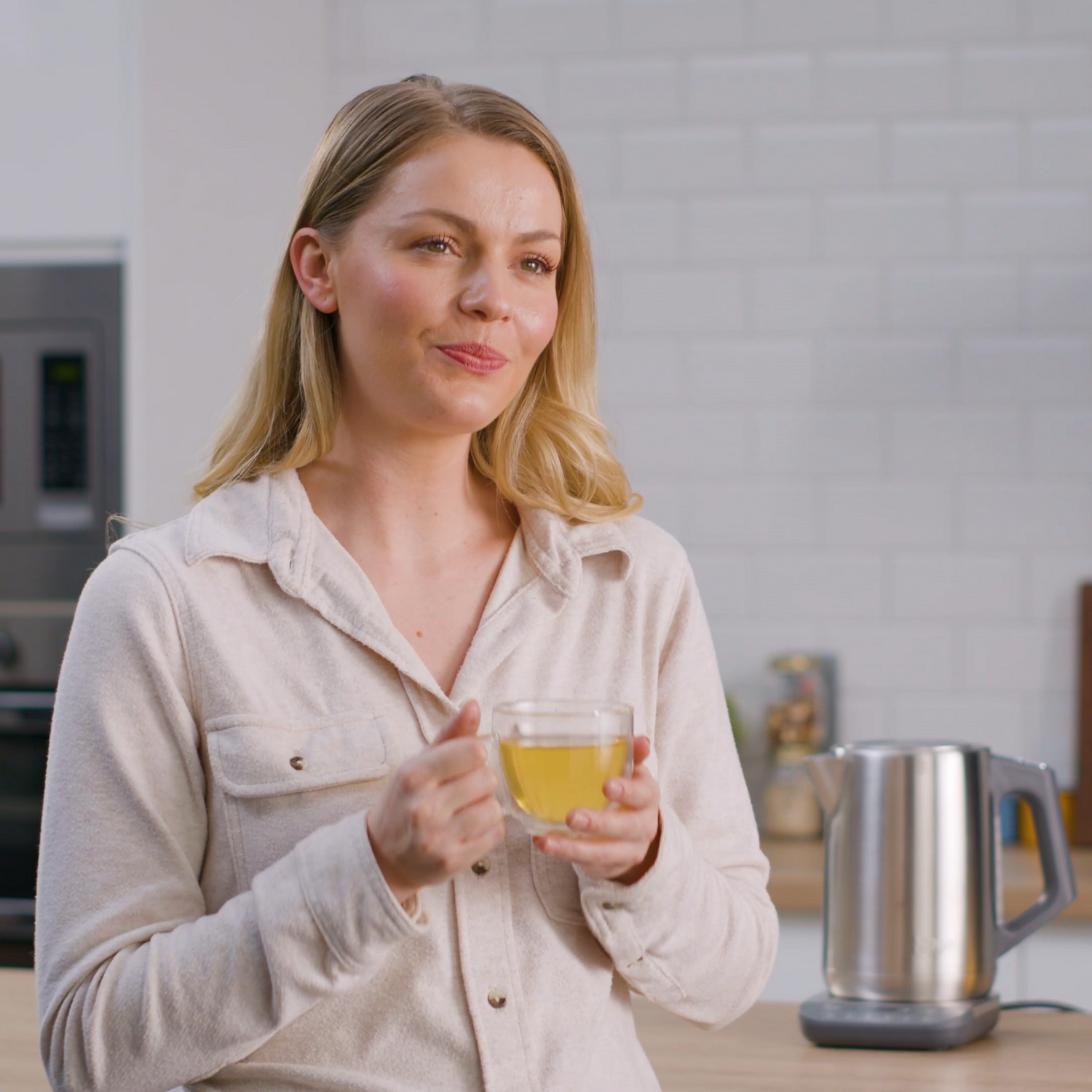 Kobieta trzymająca szklankę herbaty w kuchni, uśmiechnięta.