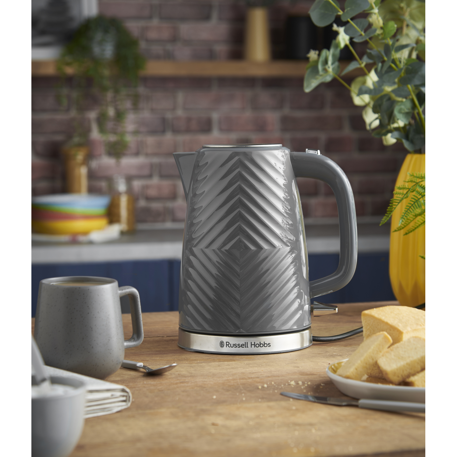 Bouilloire électrique grise avec un motif géométrique, posée sur une table en bois. Tasse et nourriture à proximité.