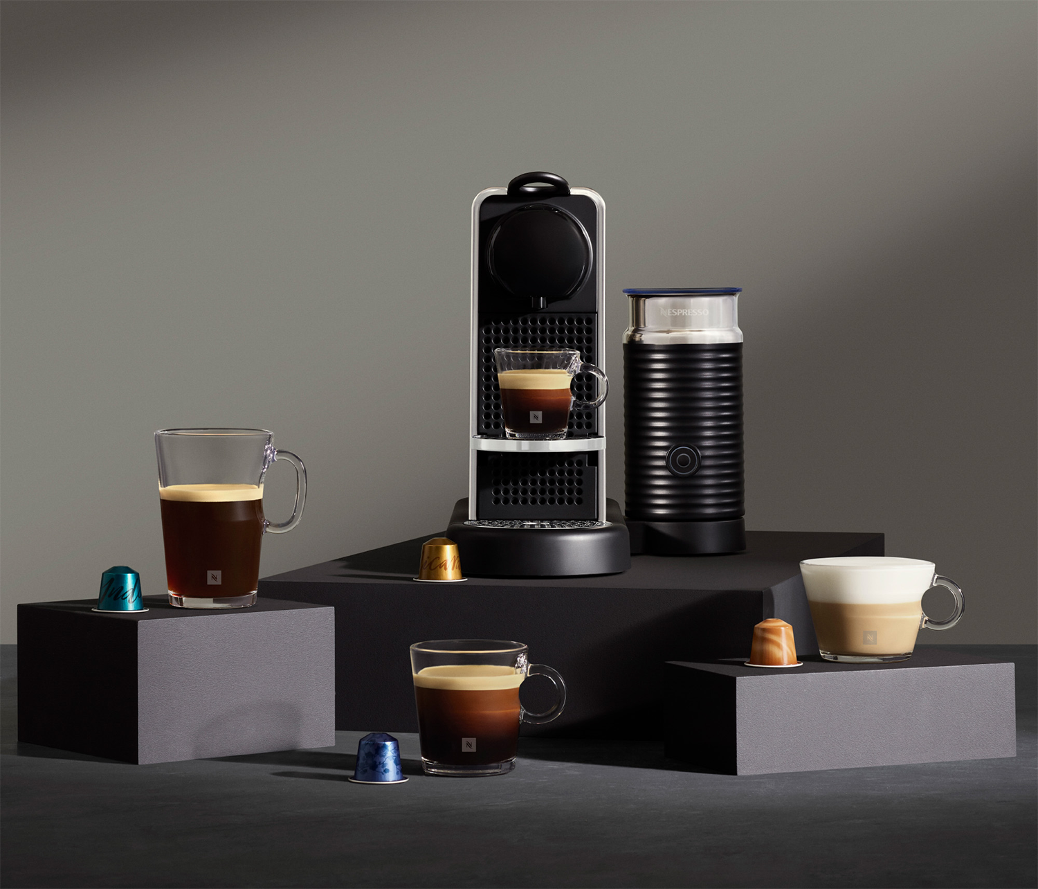Ekspres do kawy Nespresso z filiżankami, kapsułkami i spieniaczem do mleka. Na szarych platformach, szare tło.