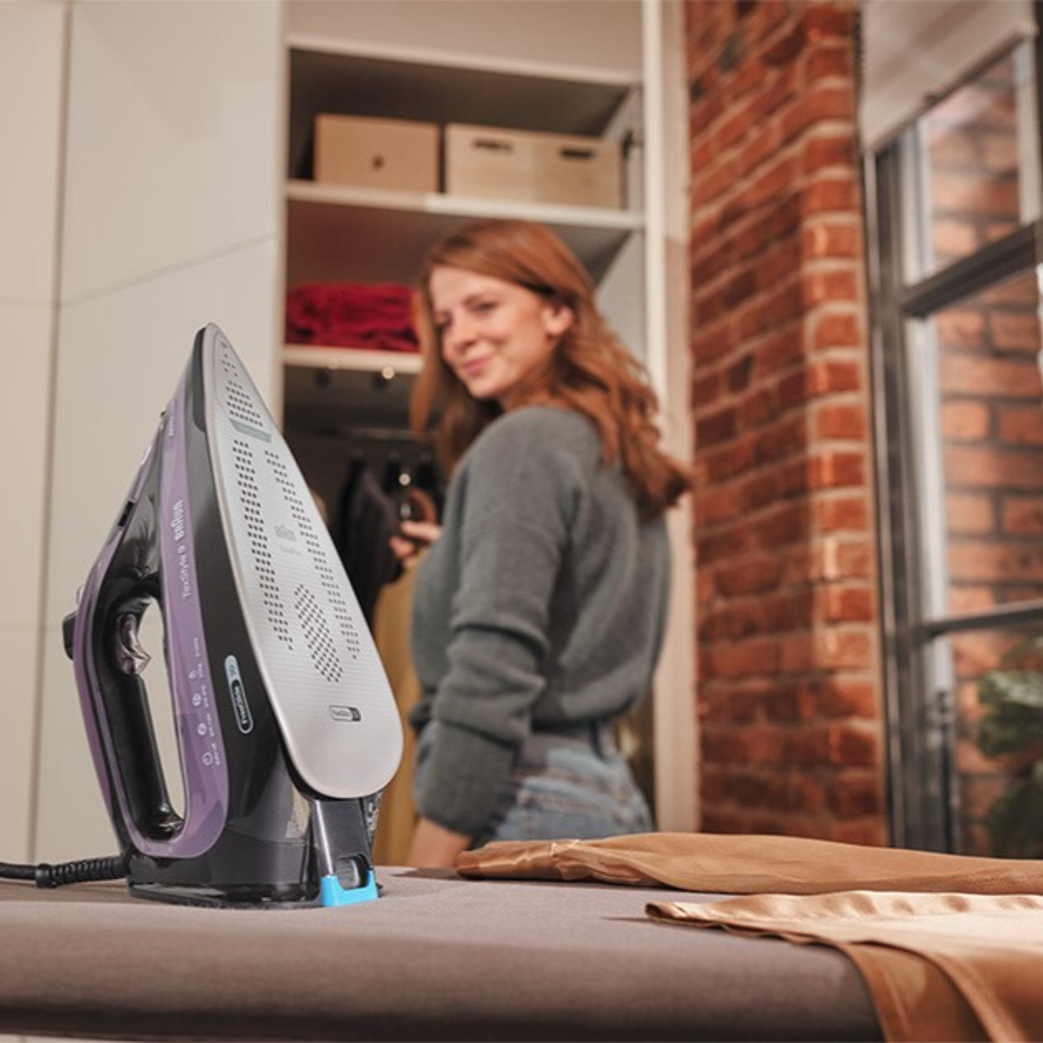 Frau bügelt mit einem schwarz-violetten Bügeleisen. Im Hintergrund ein Schrank und eine Backsteinmauer.