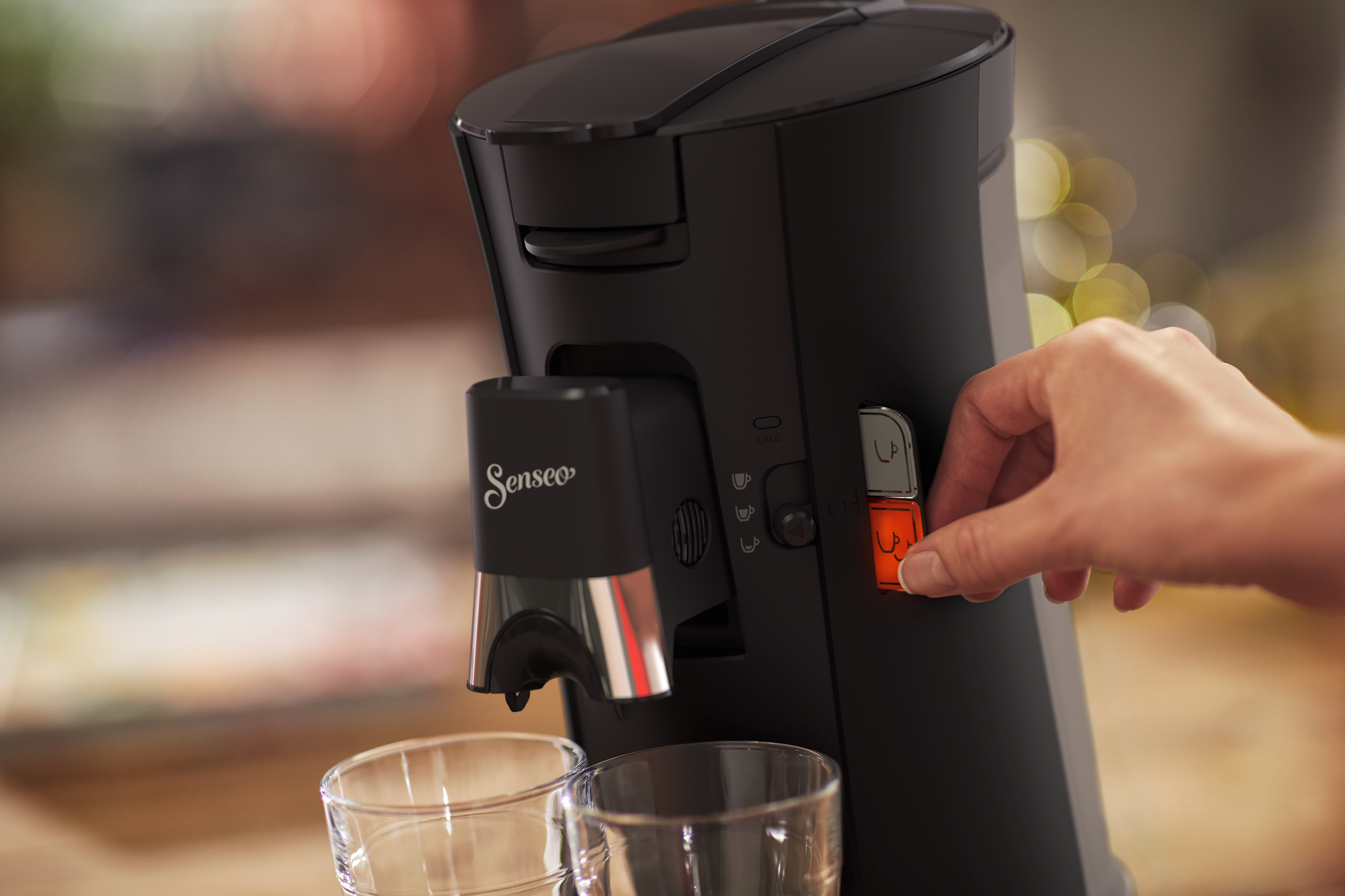 A black Senseo coffee machine with a hand turning a dial. Two glasses are in front.