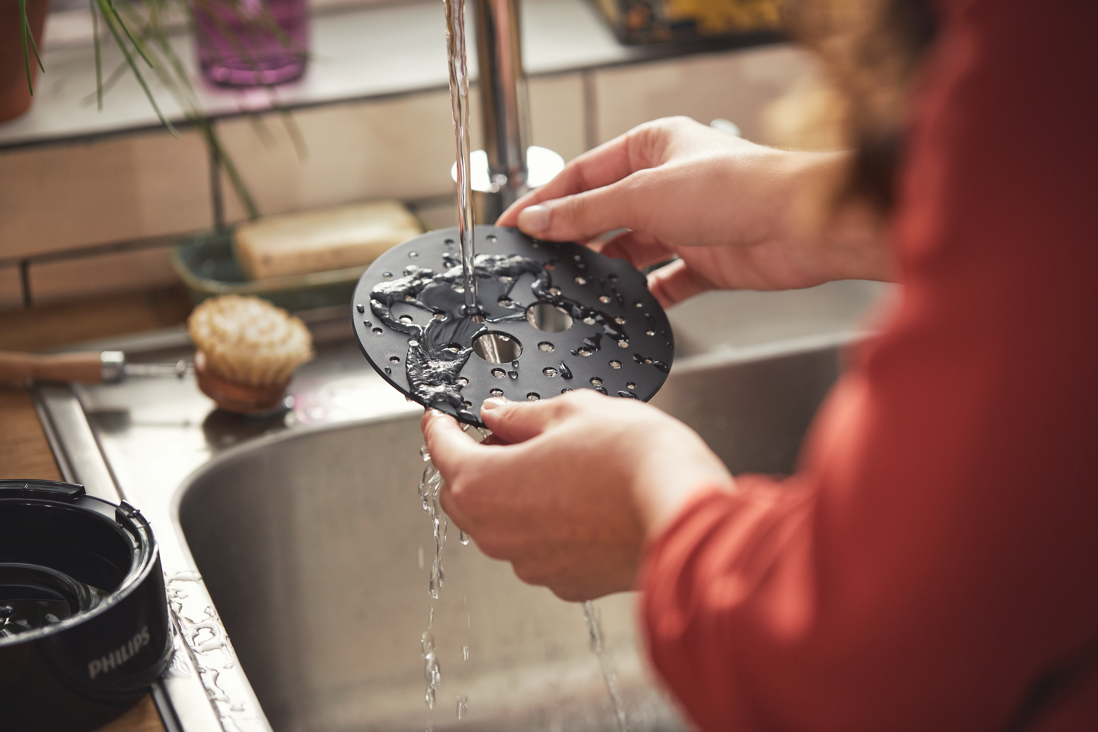 Hands washing a black, perforated air fryer accessory under running water in a sink.