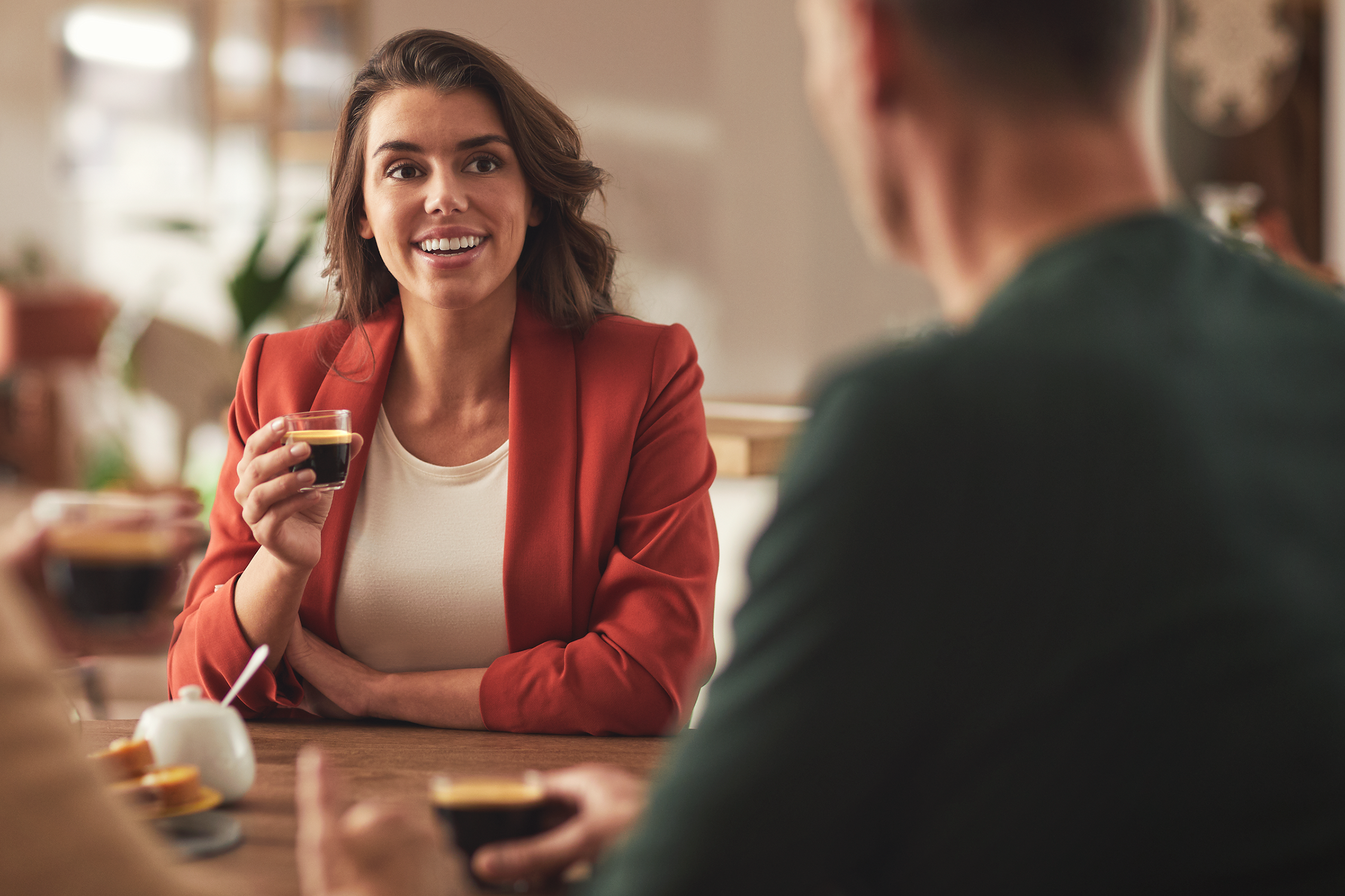 Woman holding a coffee, smiling. Another person and a table are in the scene.