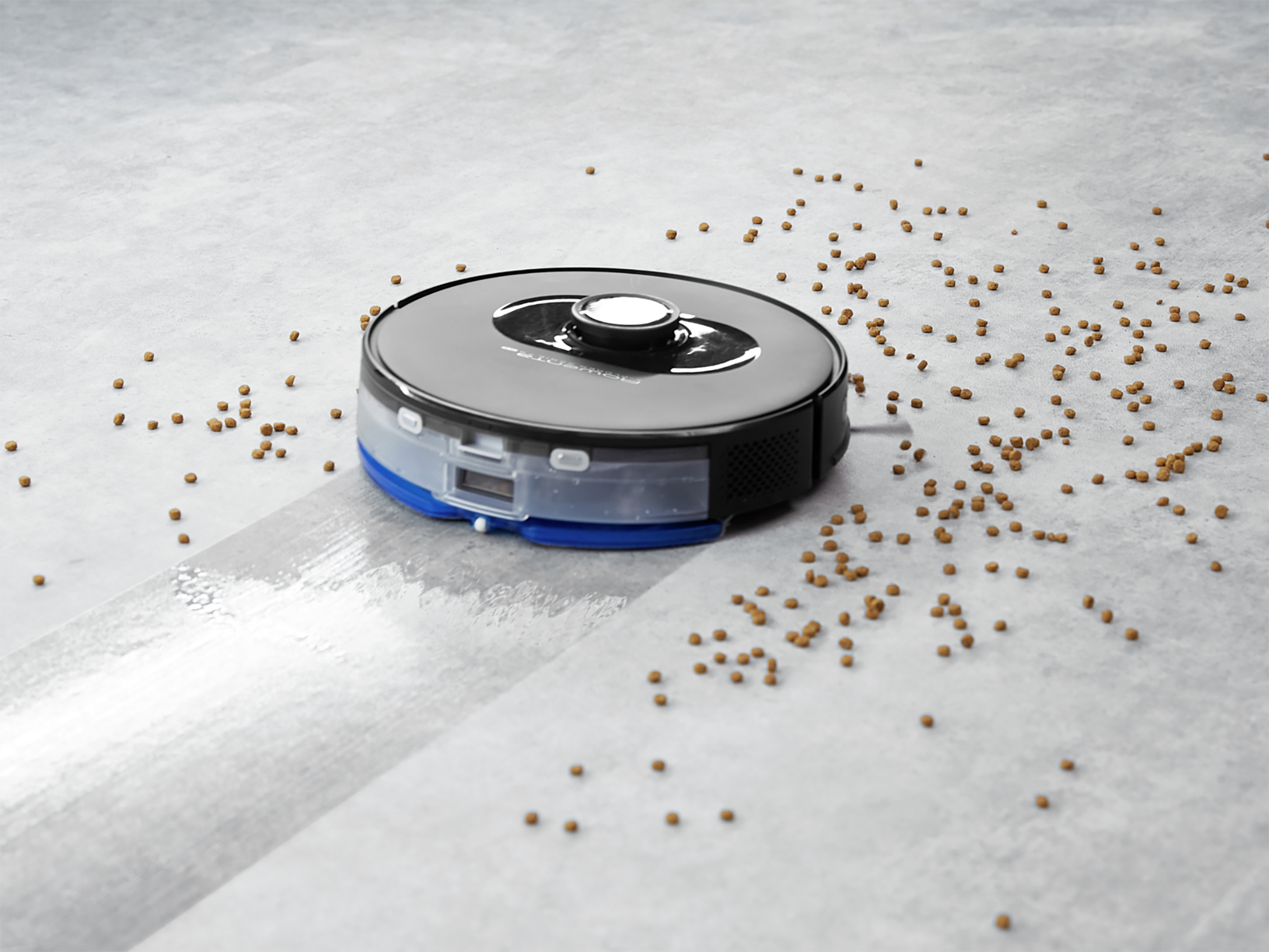 Robot aspirador limpiando un suelo gris. Bolitas marrones dispersas alrededor del aparato.