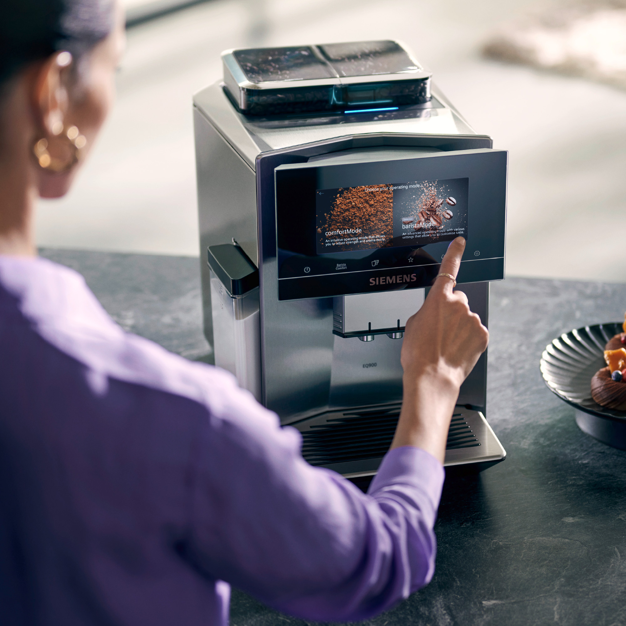 Woman using a silver coffee machine. Touchscreen, various coffee options visible.