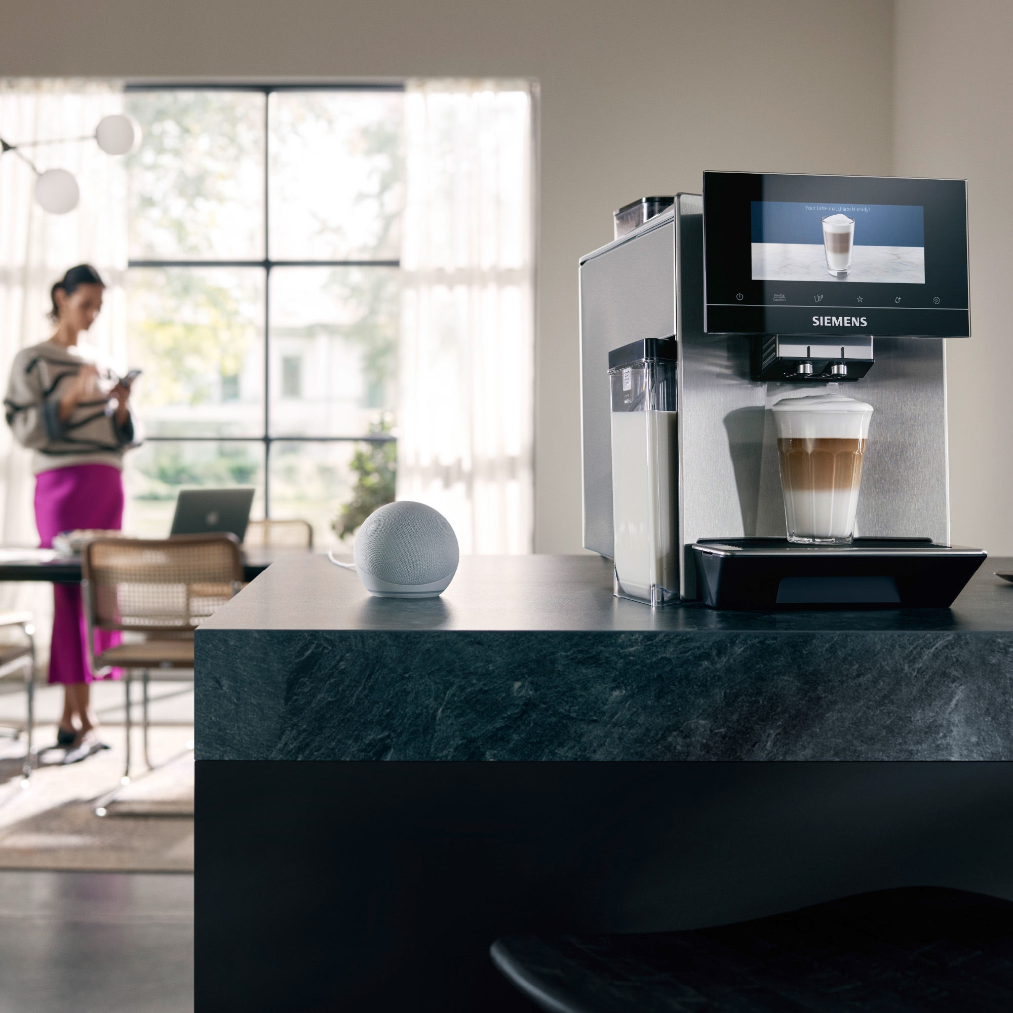 Kitchen with coffee machine, latte in cup. Woman with phone, smart speaker on counter.