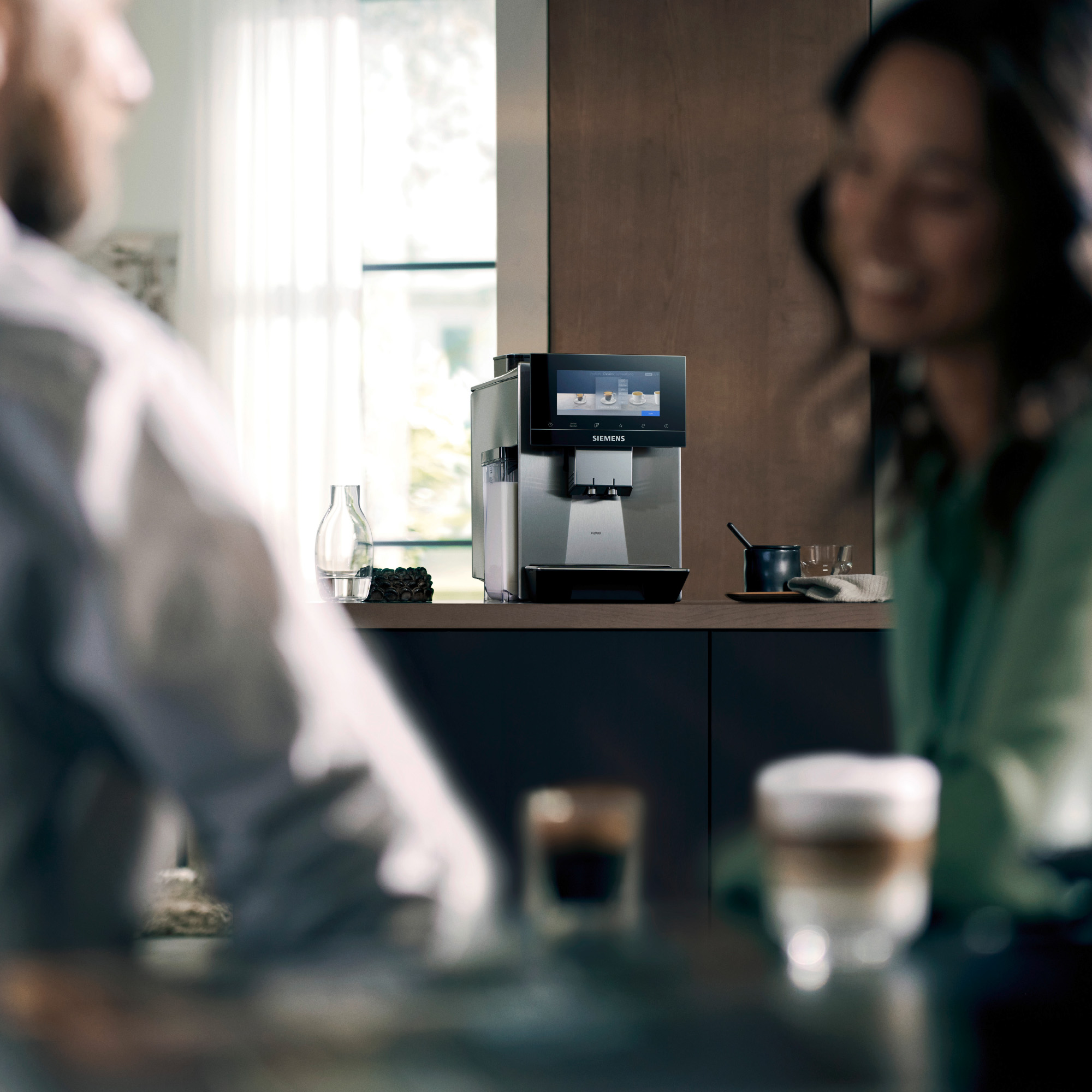 A couple in a kitchen with coffee machine. Two glasses with coffee are in focus.