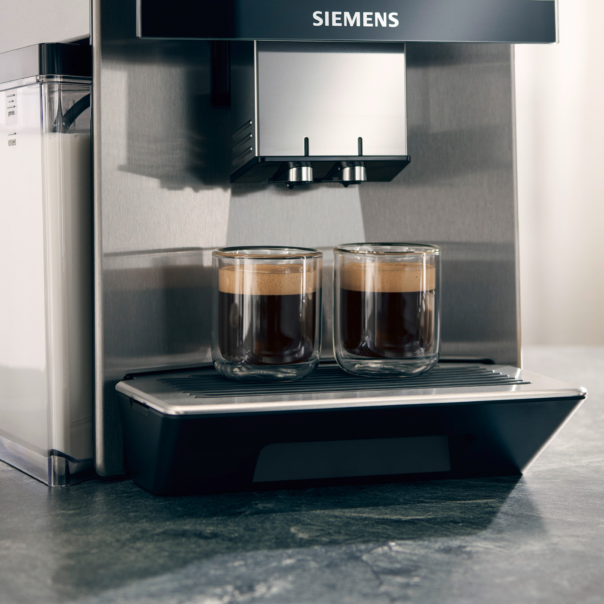 A silver Siemens coffee machine with two glasses filled with coffee. The machine is on a dark gray countertop.