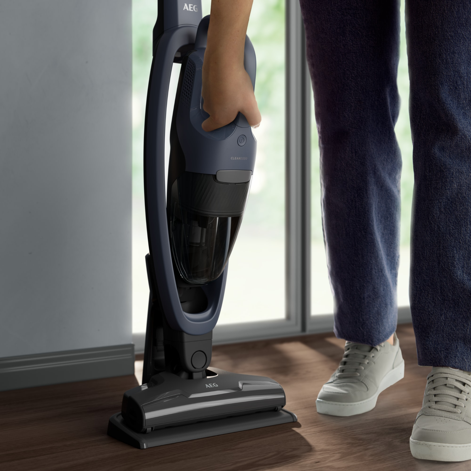 Person holding a dark blue cordless vacuum cleaner. Wooden floor, white wall, and natural light.