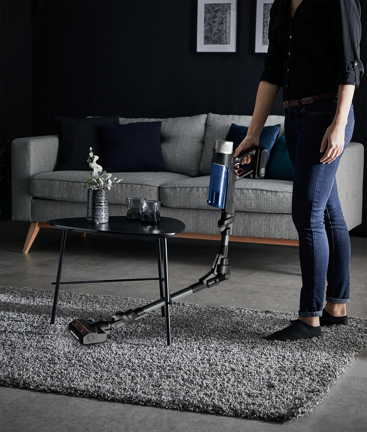 Femme passant l'aspirateur sur un tapis avec un aspirateur sans fil, canapé et table d'appoint en arrière-plan.
