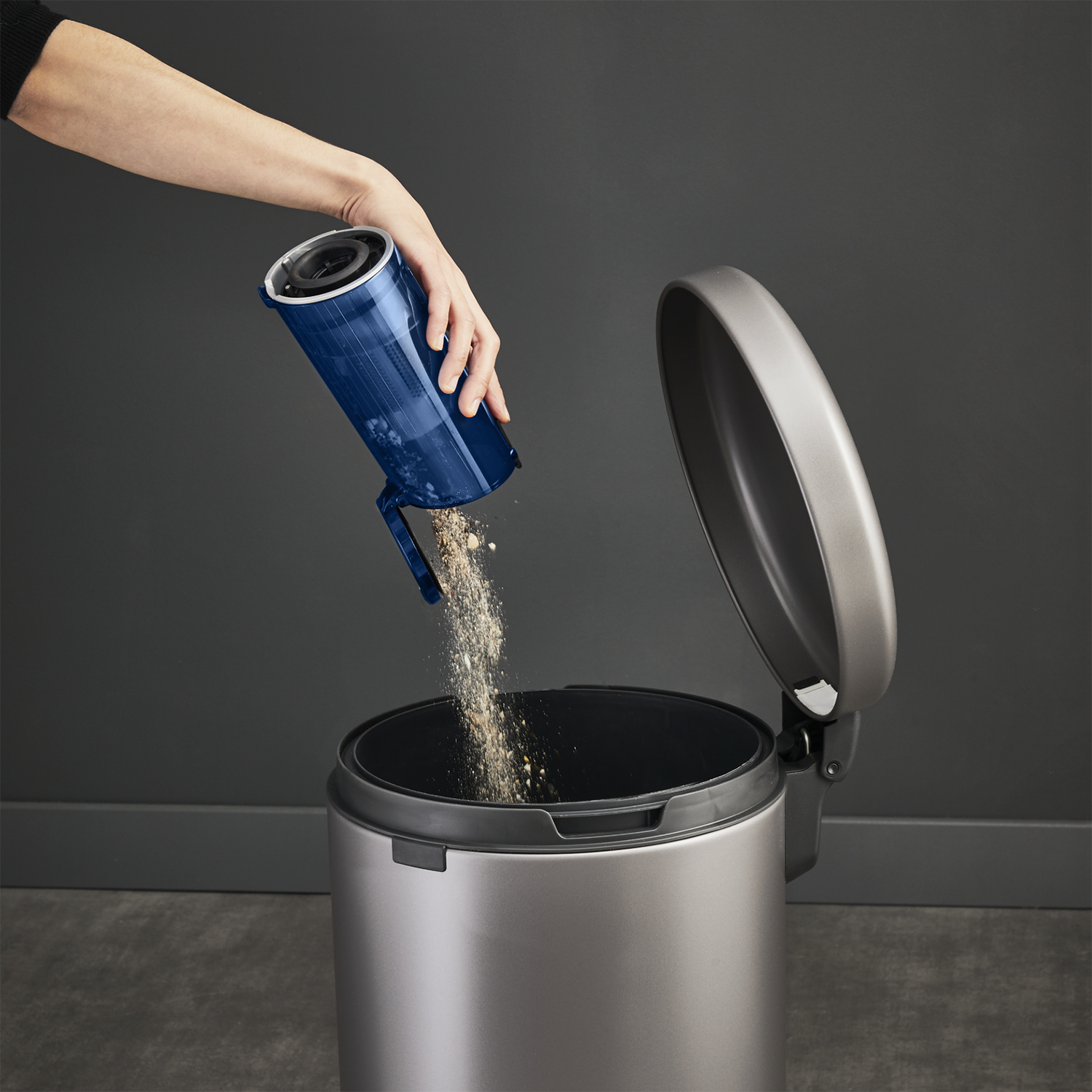 Une main vide un récipient bleu d'aspirateur dans une poubelle argentée.