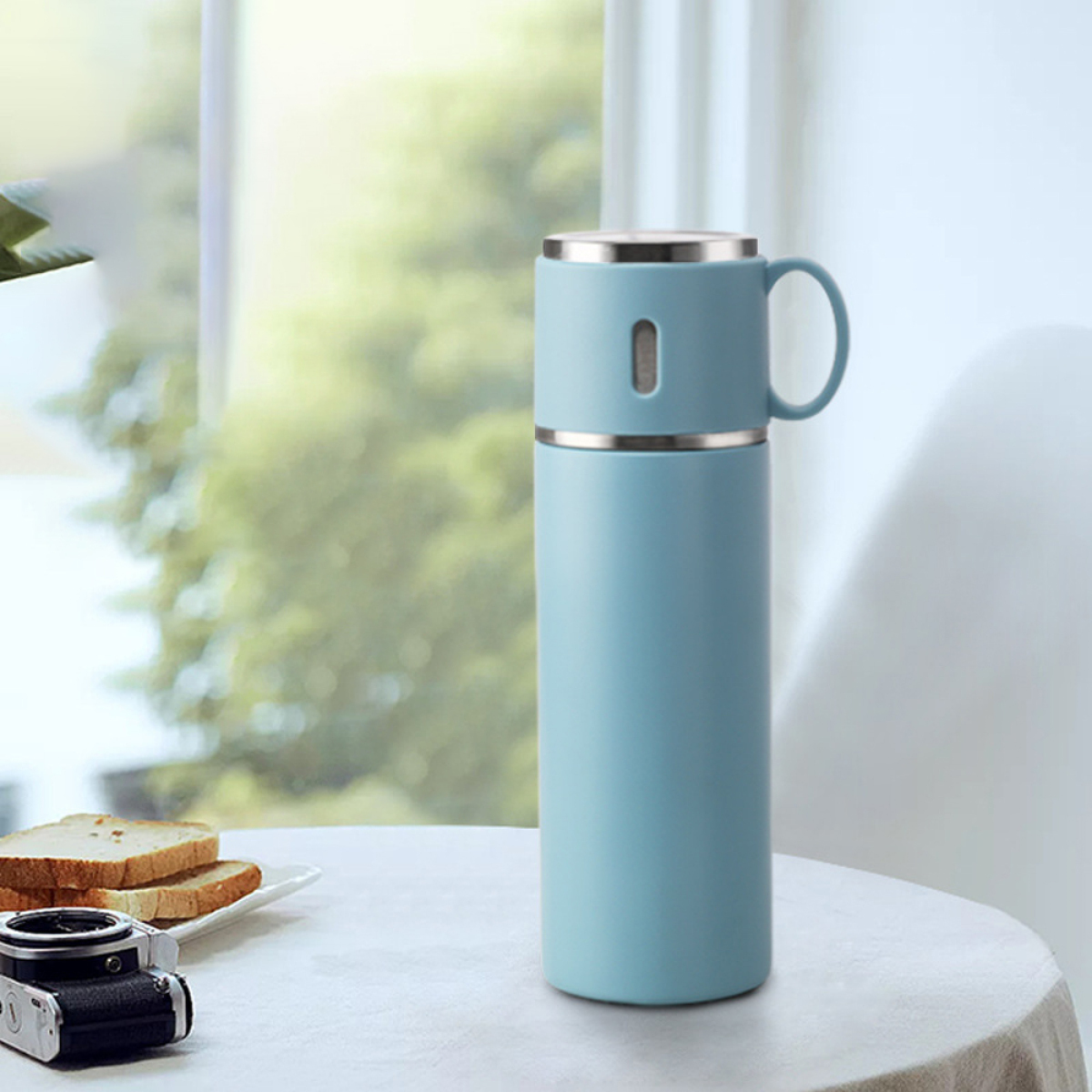 Blaue Thermoskanne mit Tasse, neben Brotscheiben und einer Kamera auf einem Tisch in Fensternähe.