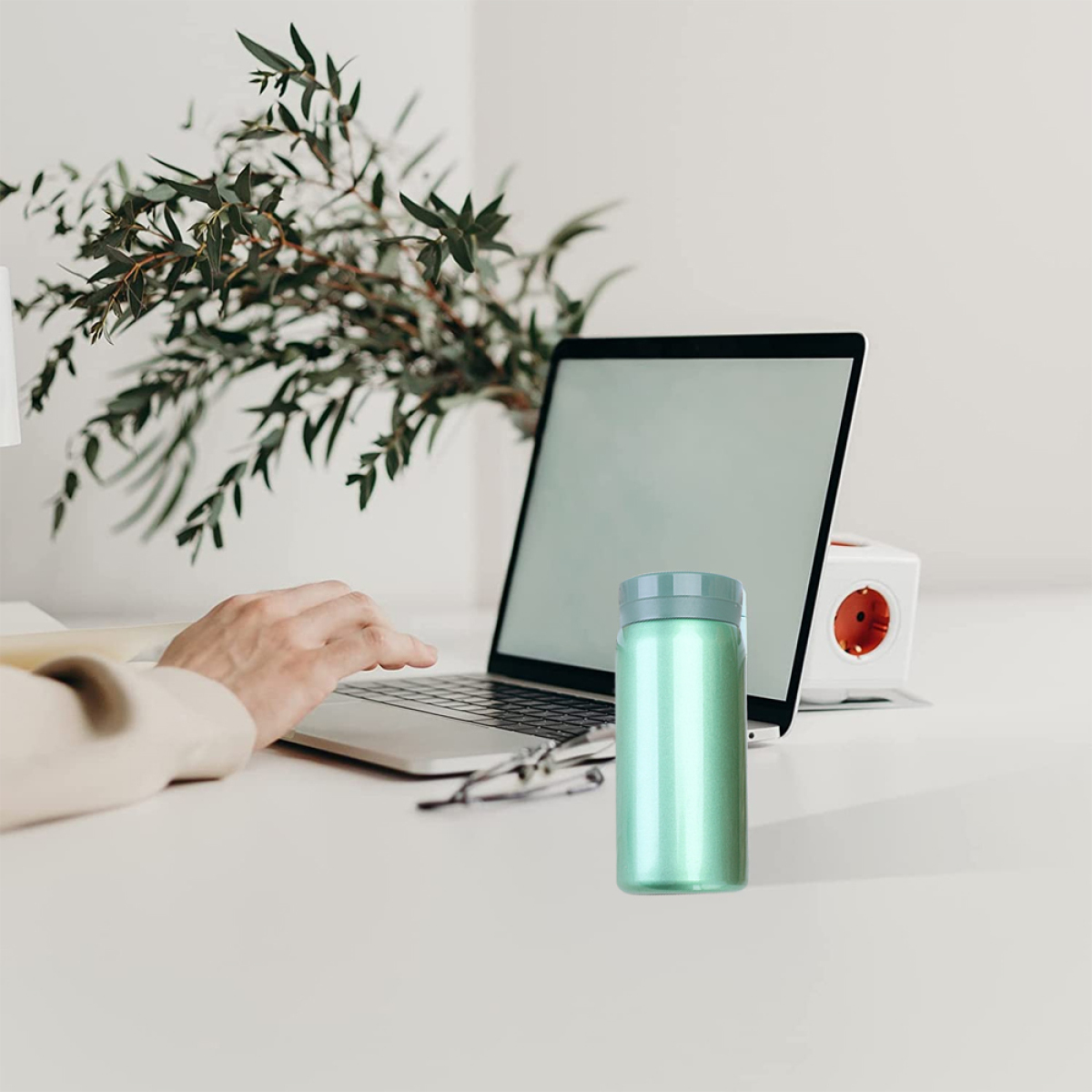 Eine grüne Reiseflasche auf einem weißen Schreibtisch neben einem Laptop und einer grünen Pflanze.
