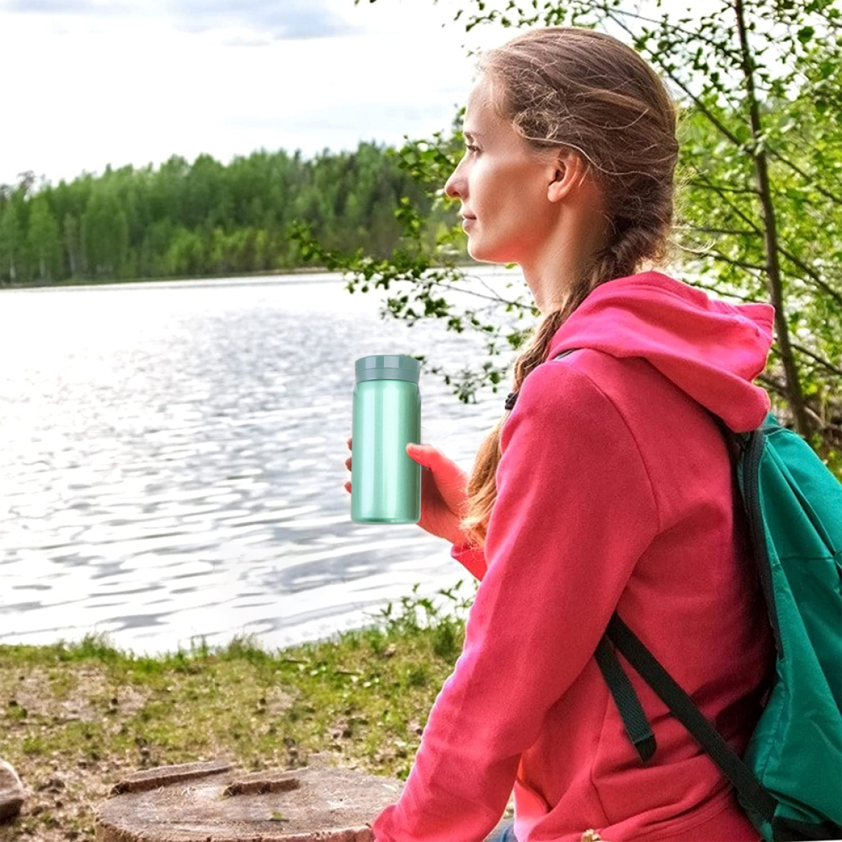 Eine Frau in einem rosa Hoodie sitzt an einem See. Sie hält einen grünen Becher. Hintergrund: See und grüne Bäume.