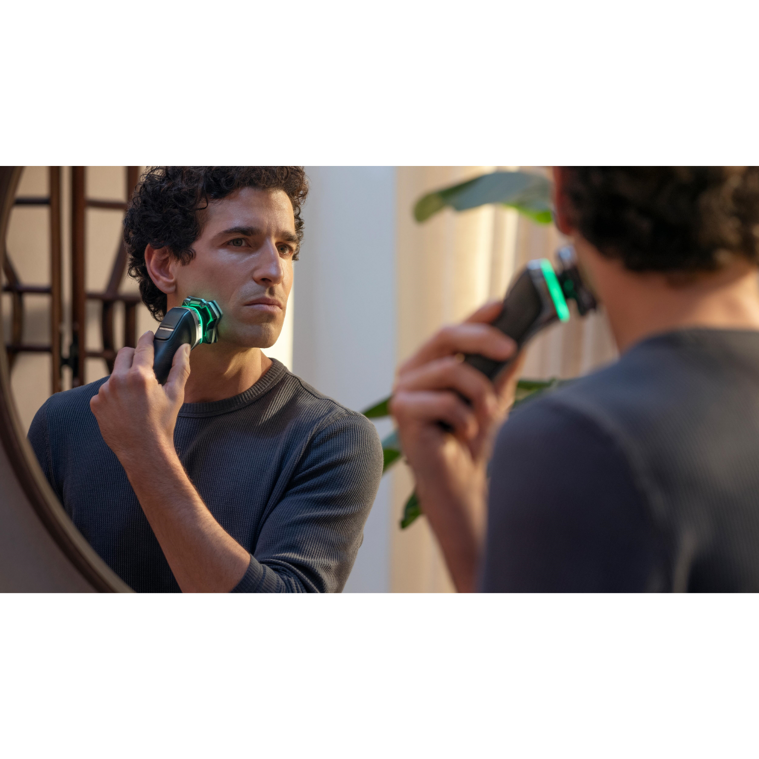 Mężczyzna goli się w lustrze. Goli twarz czarno-zieloną golarką elektryczną.