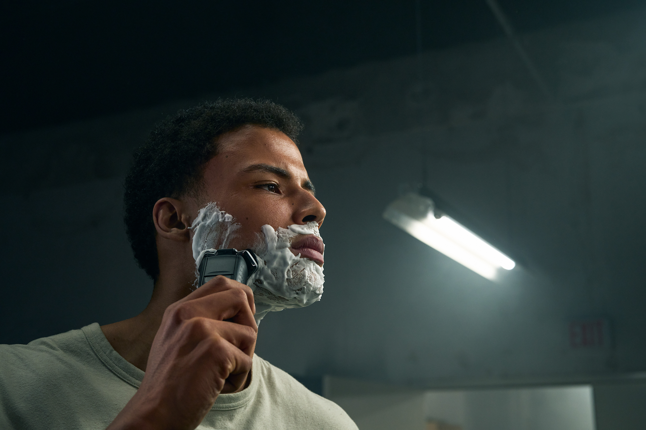 Mężczyzna goli się czarną golarką elektryczną. Pianka na twarzy, światło z góry, ciemne tło.