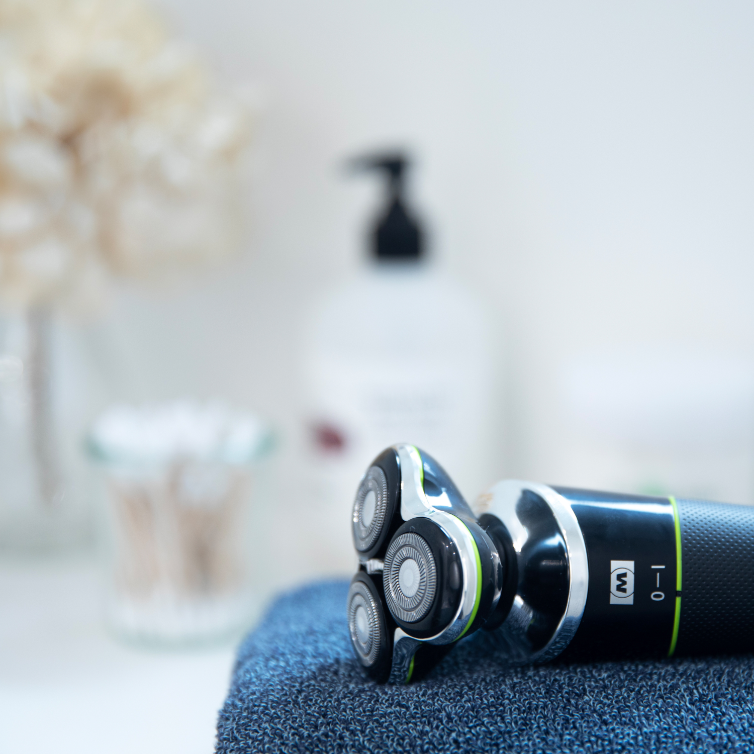 Golarka elektryczna na niebieskim ręczniku, z butelką i patyczkami kosmetycznymi w tle.