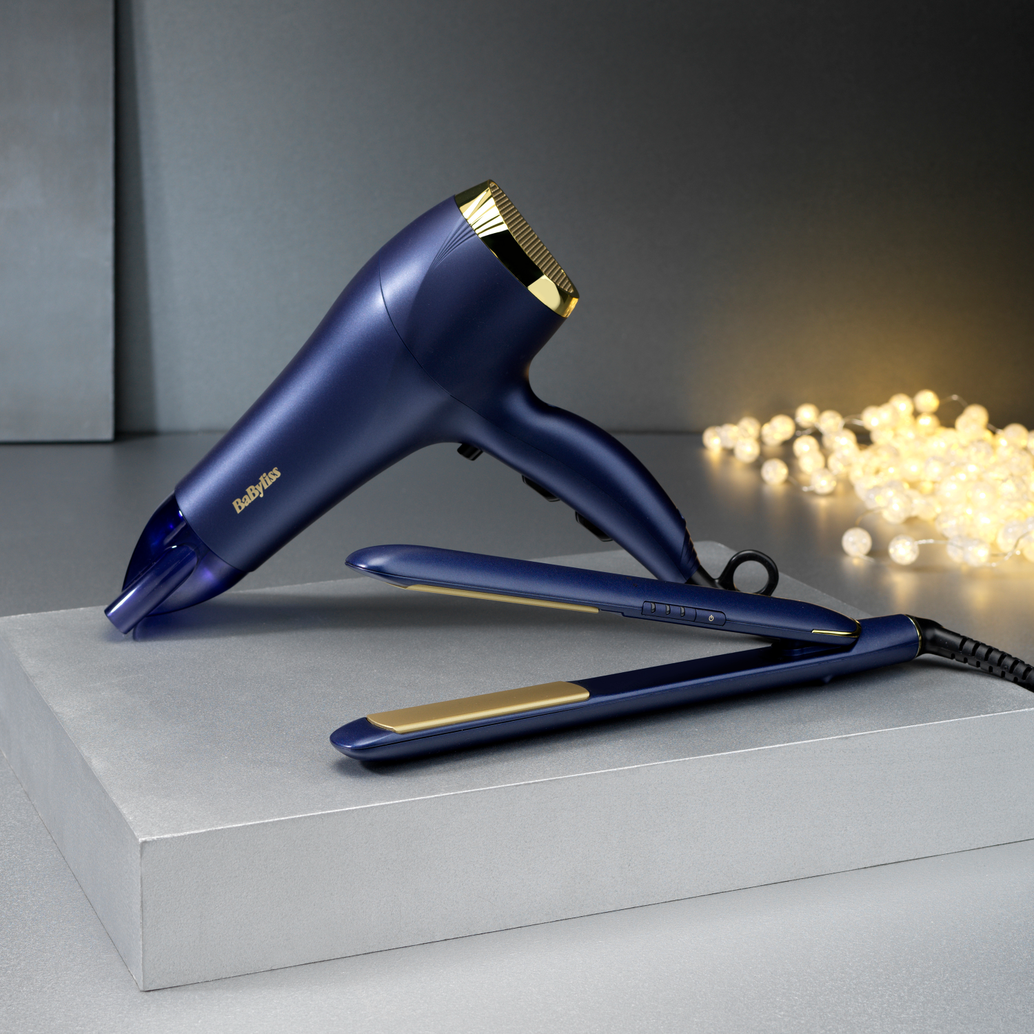 Niebieska i złota suszarka do włosów i prostownica na szarej powierzchni. W tle sznur światełek.