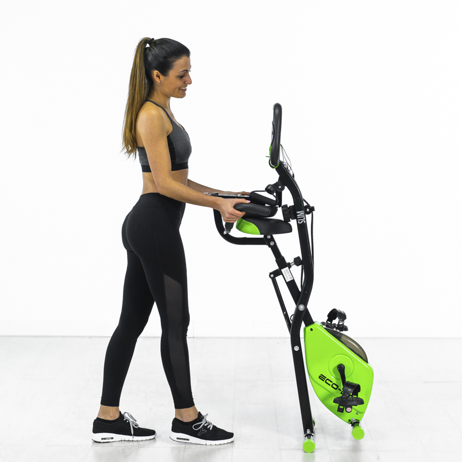 Mujer de pie junto a una bicicleta estática verde y negra. Lleva leggings negros y un sujetador deportivo.