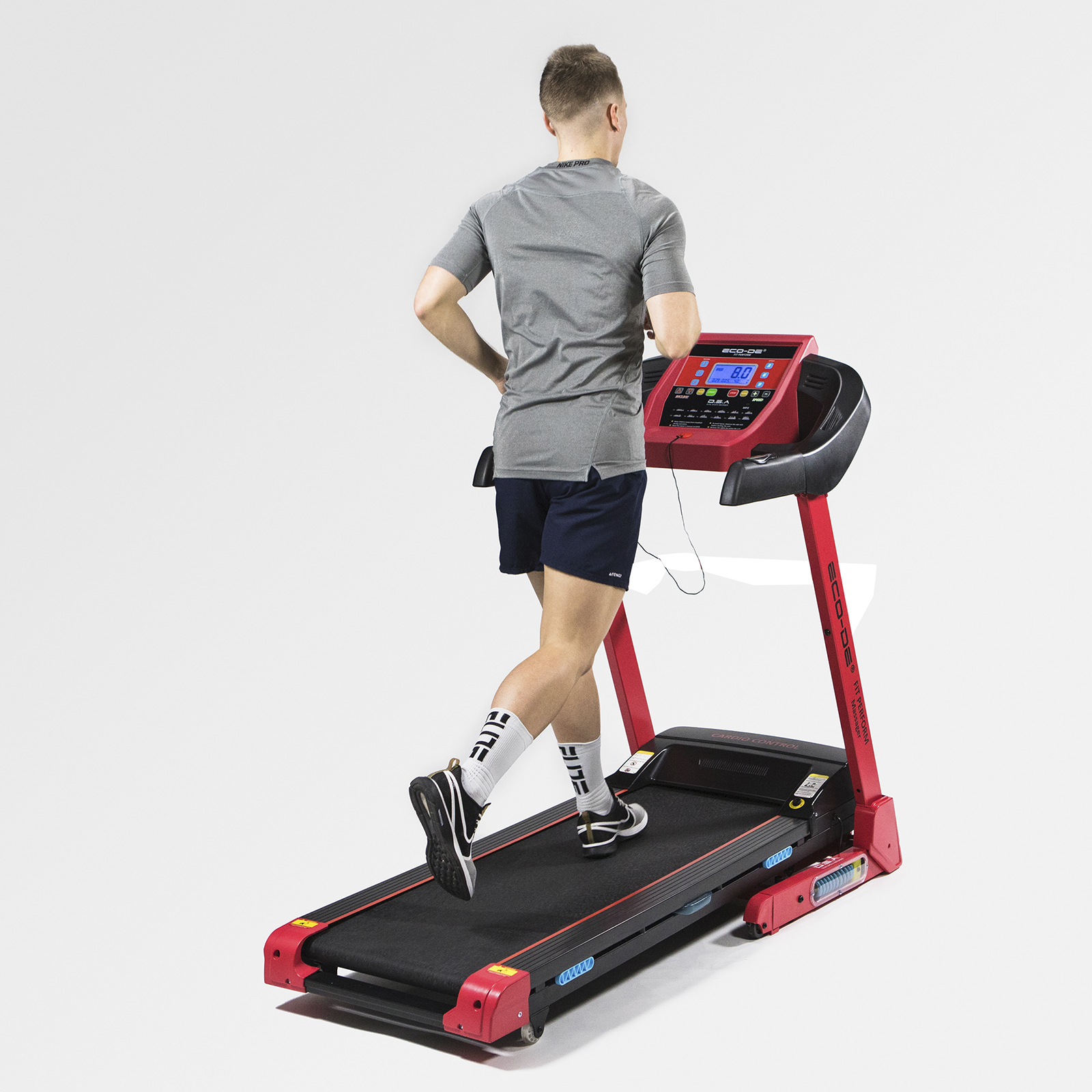 Un hombre corre en una cinta de correr roja. Lleva una camisa gris, pantalones cortos oscuros y calcetines blancos.