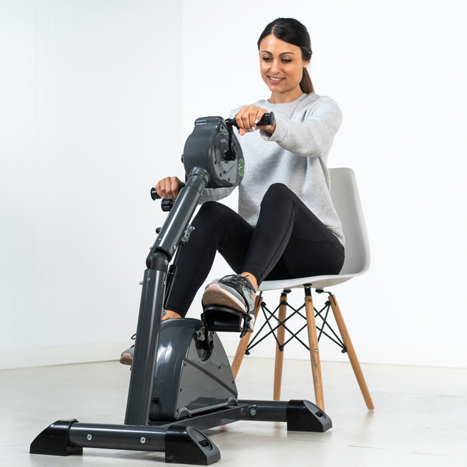 Mujer ejercitándose con bicicleta estática portátil en una silla, fondo blanco.