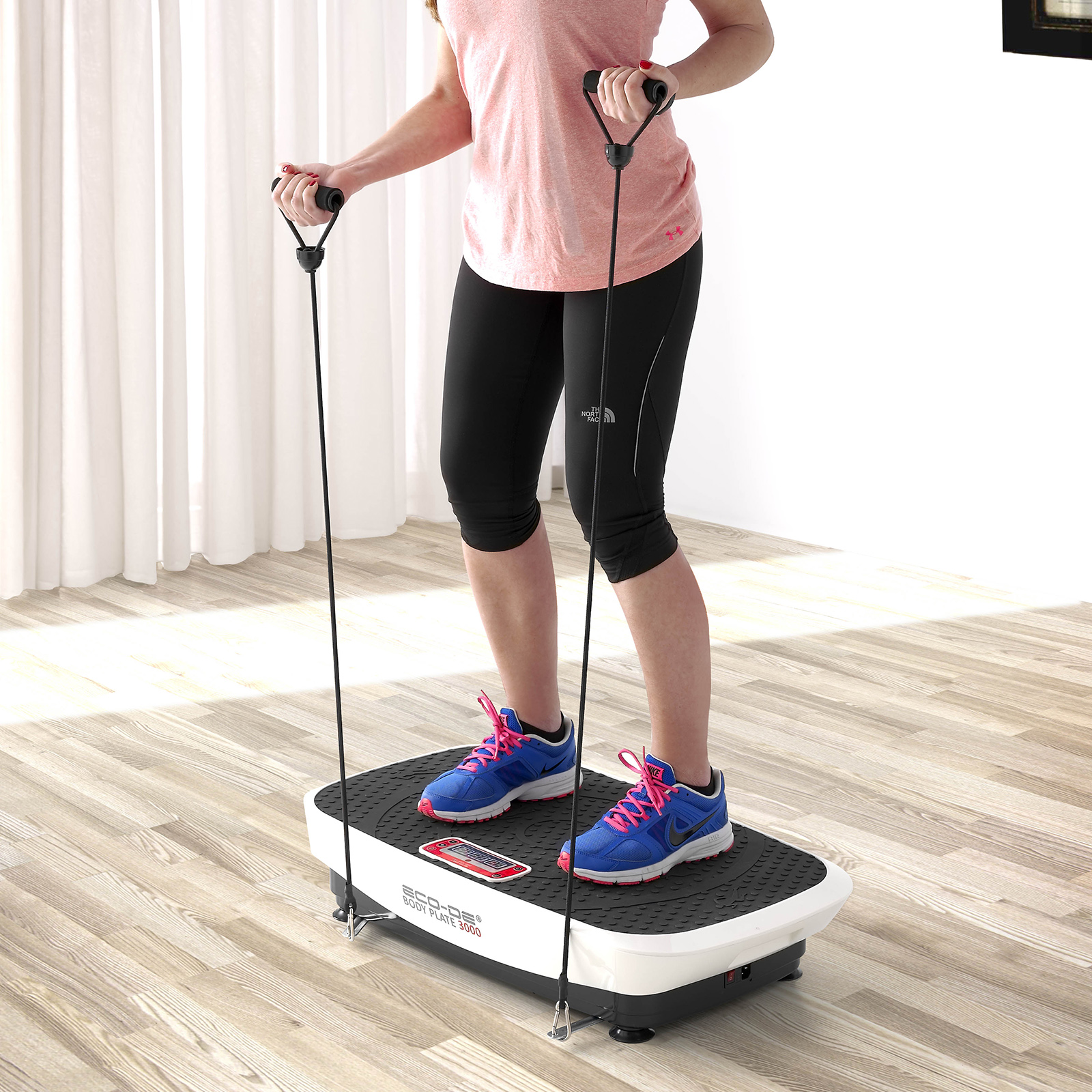 Mujer en plataforma vibratoria, sosteniendo bandas de resistencia, top rosa, leggings negros, piso de madera.