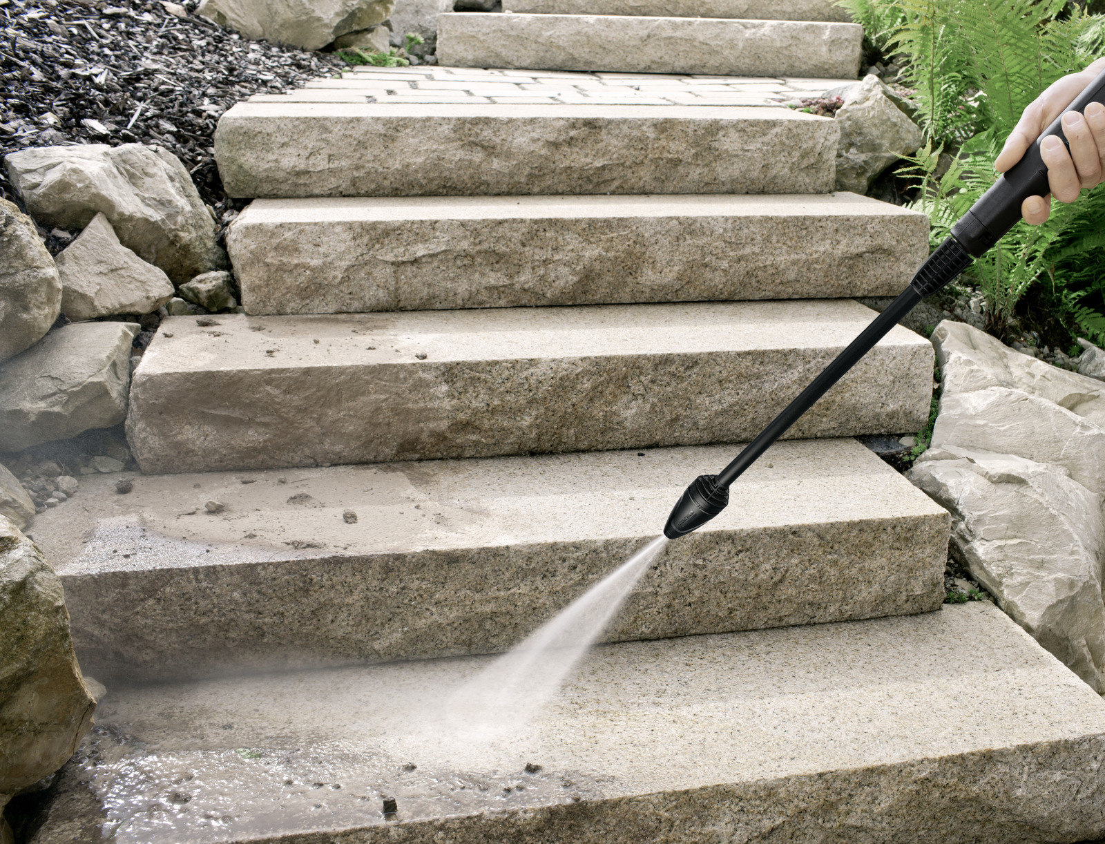 Persona che pulisce gradini di pietra con idropulitrice. Spruzzo d'acqua visibile. Sfondo: scala in pietra e vegetazione.
