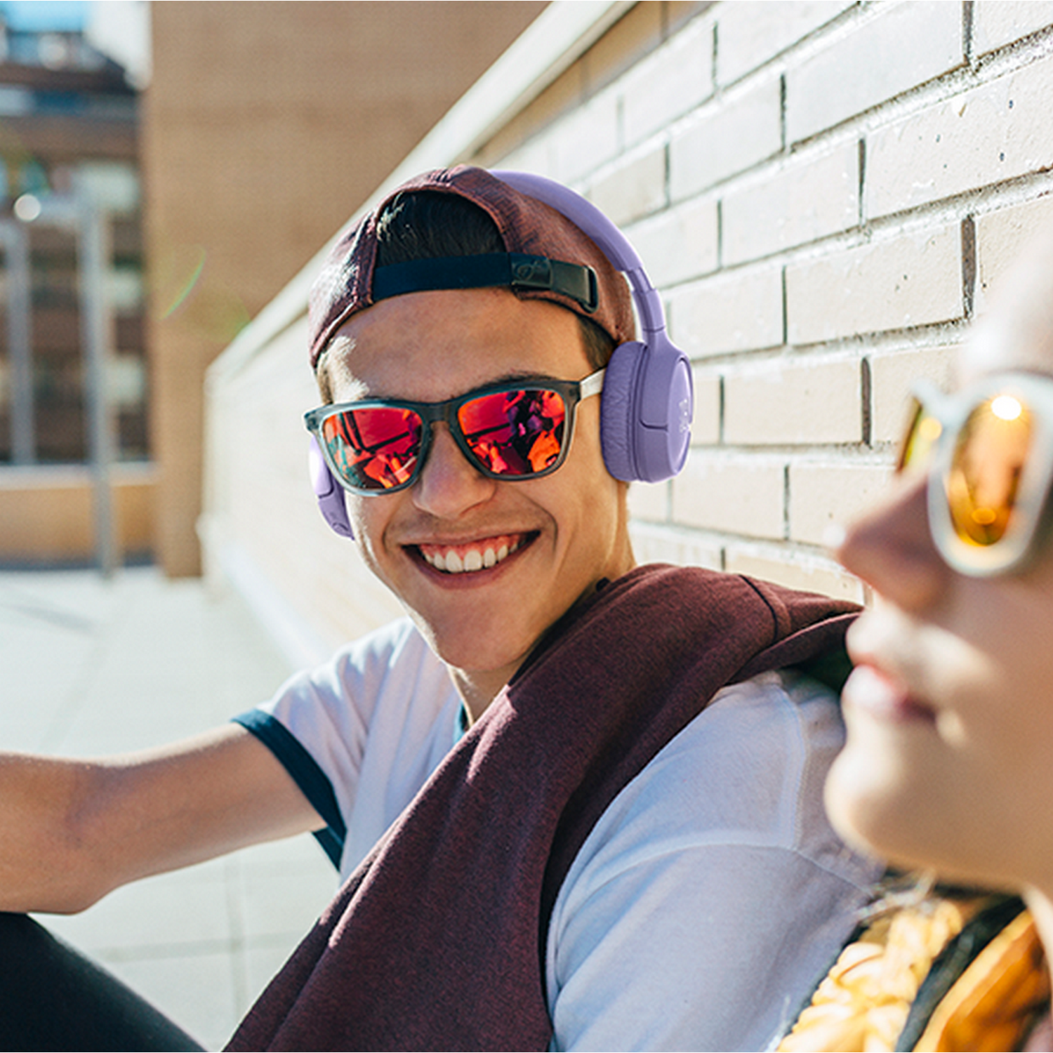 Ein junger Mann mit Sonnenbrille und lila Kopfhörern lächelt. Eine andere Person ist im Hintergrund.