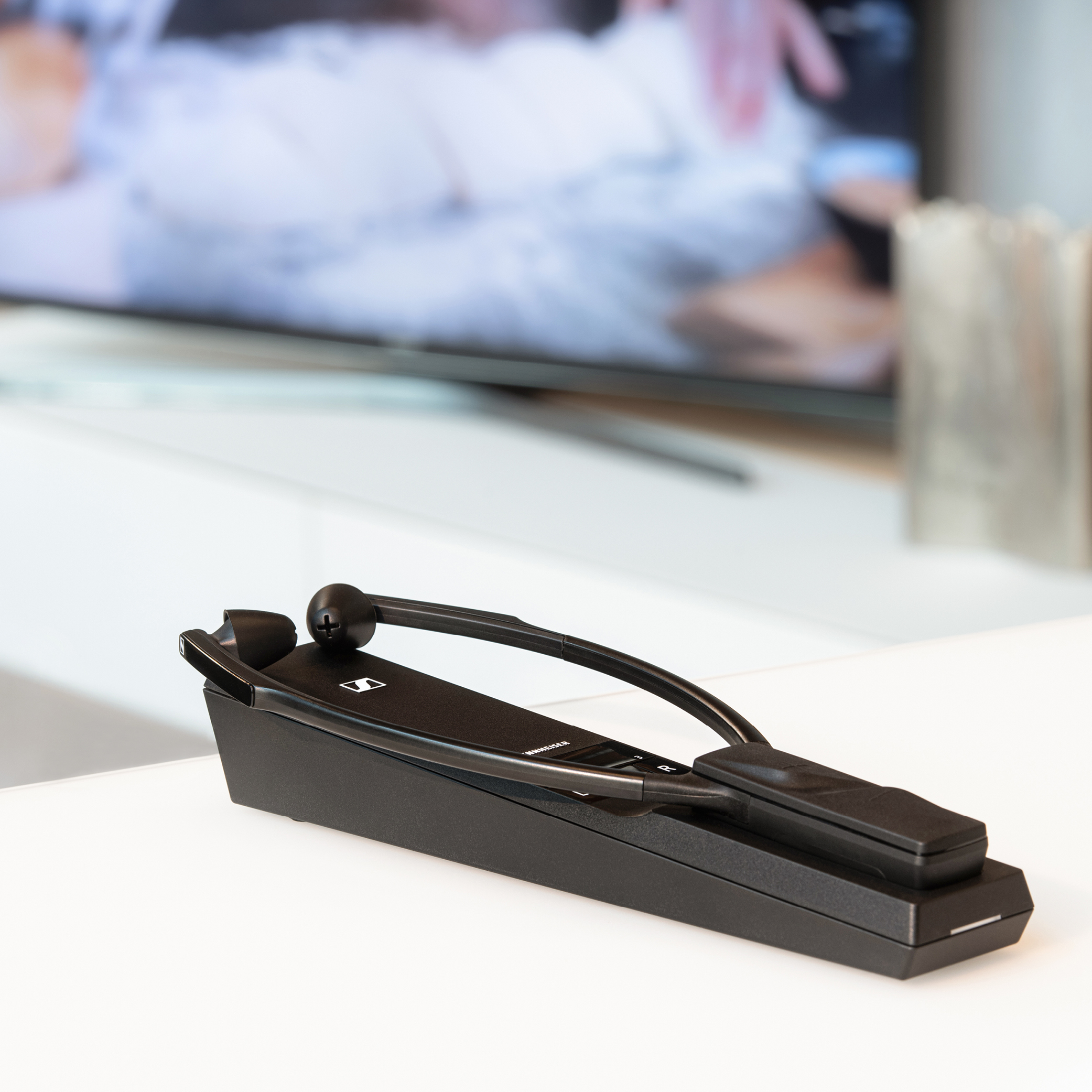 Schwarzes Hörgerät auf weißer Oberfläche, verschwommener Fernseher im Hintergrund.