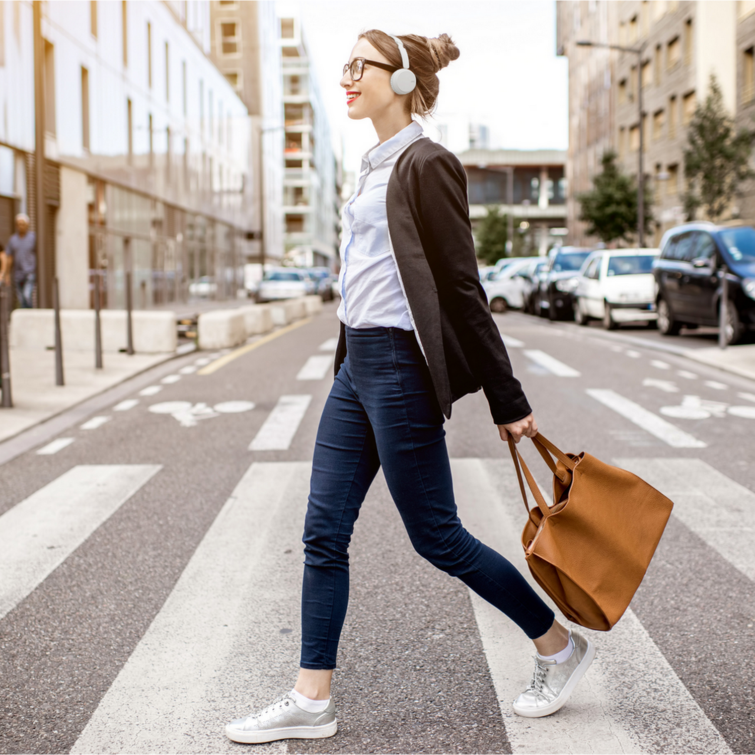 Kobieta w okularach przechodzi przez ulicę. Ma słuchawki i torbę. Budynki miejskie w tle.