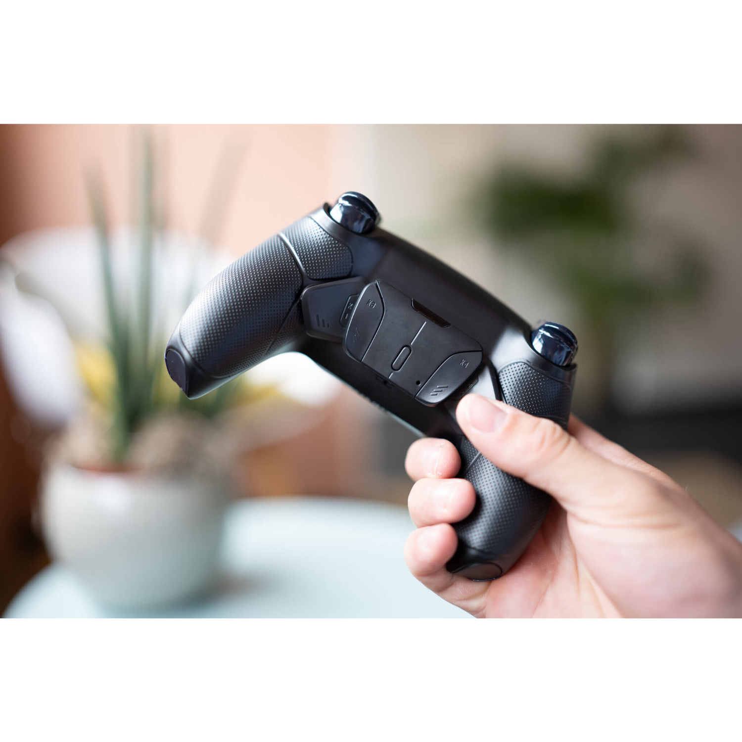Un controller nero di PlayStation 5 tenuto da una persona. Lo sfondo è sfocato con una pianta e una finestra.