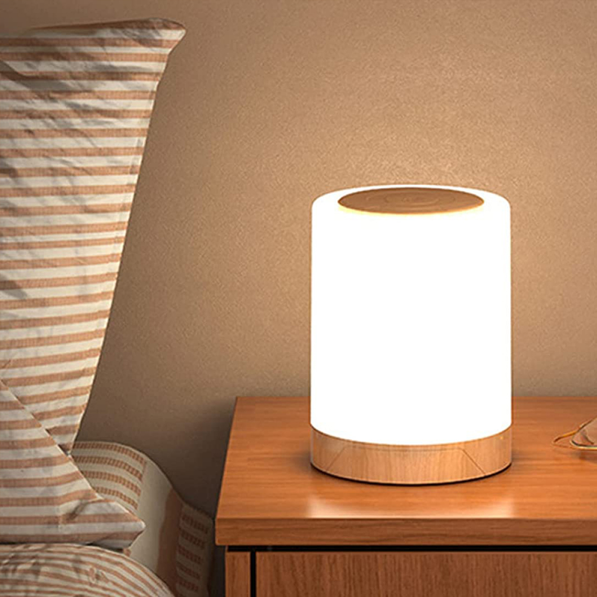 Eine beleuchtete zylindrische Lampe mit Holzfuß steht auf einem Holztisch. Im Hintergrund ist ein Bett.
