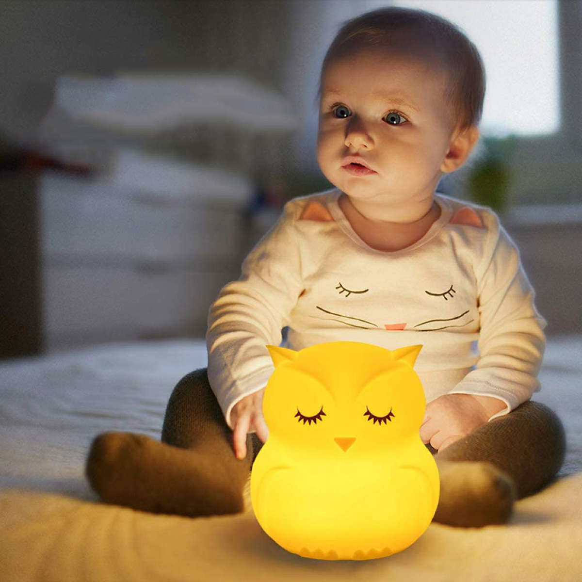 Ein Baby sitzt auf einem Bett neben einem gelben eulenartigen Nachtlicht. Das Baby schaut etwas an.