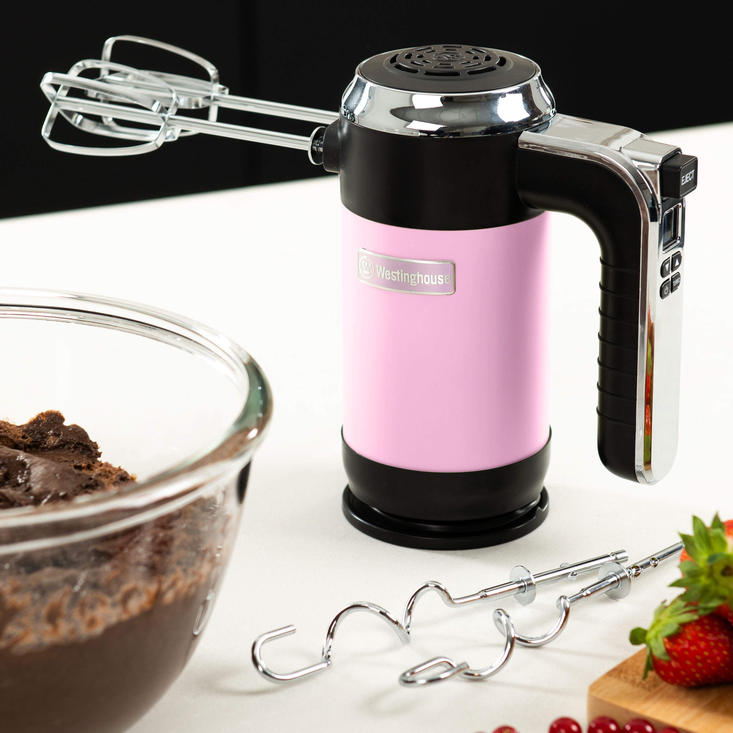 Pinker und schwarzer Handmixer mit Zubehör auf weißer Oberfläche. Eine Schüssel mit Schokolade und Erdbeere.