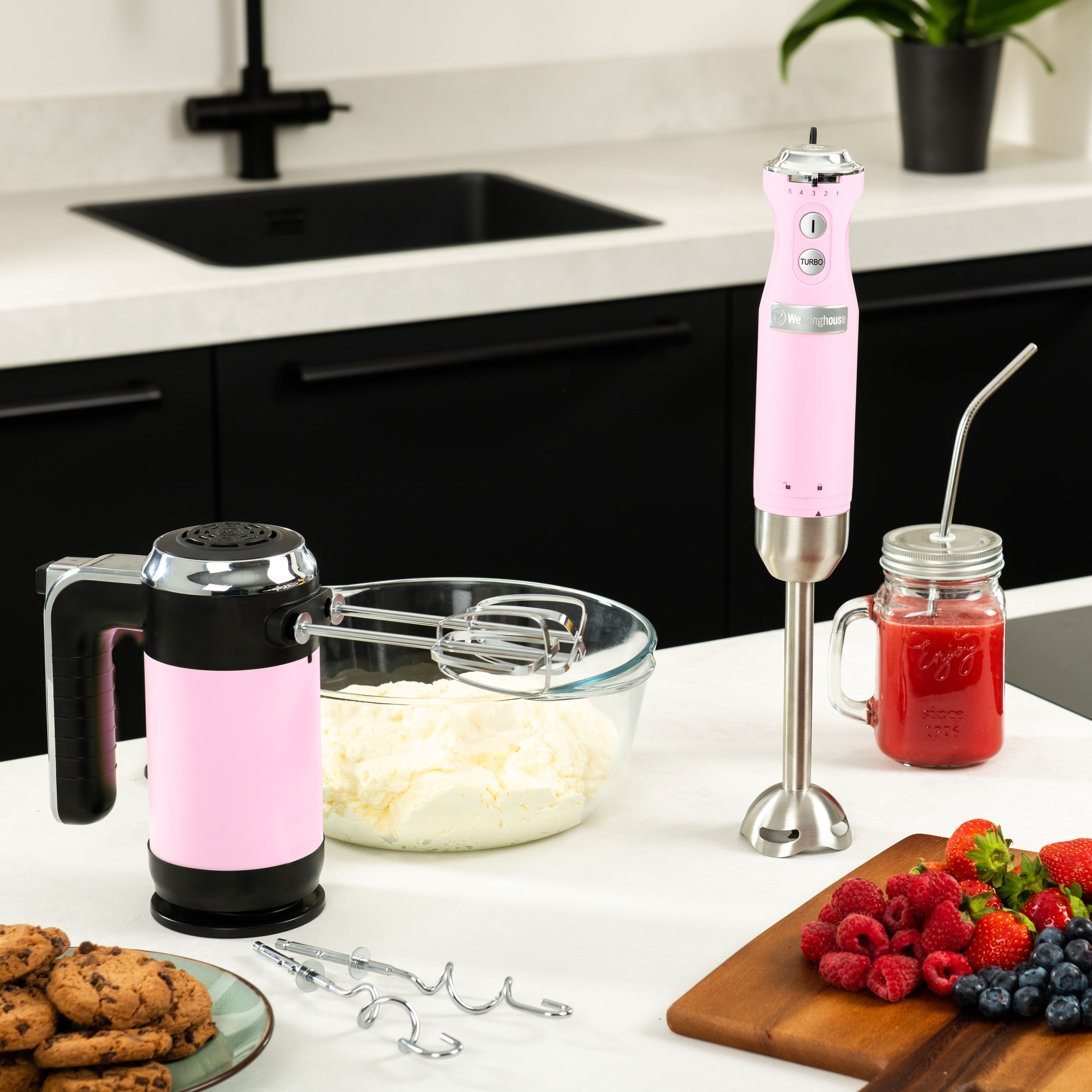 Rosa Stabmixer und Mixer auf weißer Oberfläche, mit Lebensmitteln und Zutaten drumherum.
