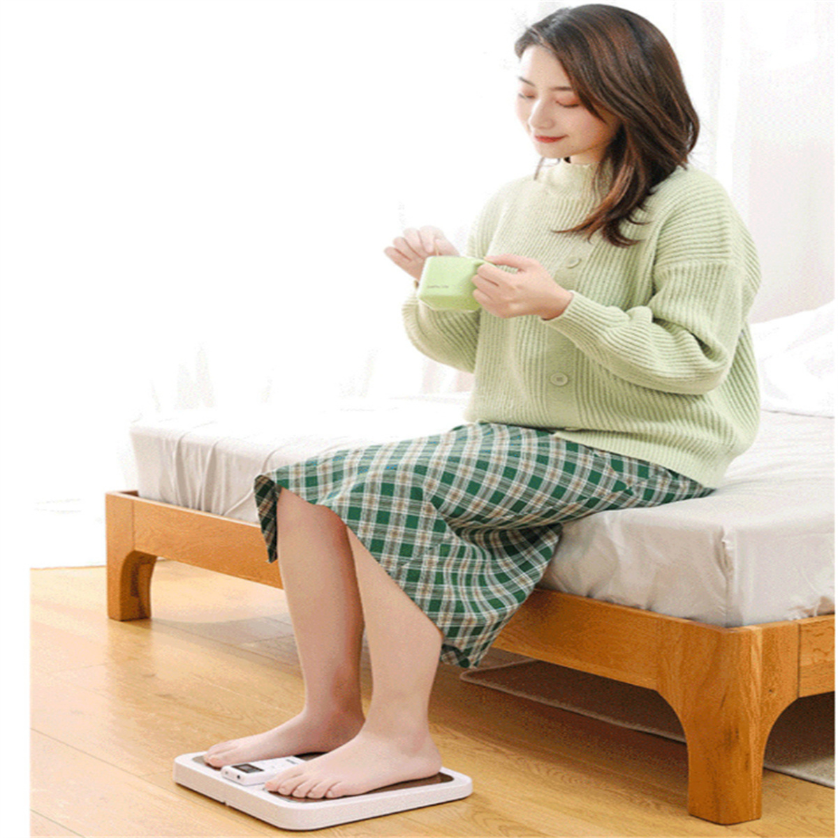Eine Frau sitzt auf einem Bett mit einer Waage unter ihren Füßen und hält einen Becher. Sie trägt einen Pullover und karierte Hose.