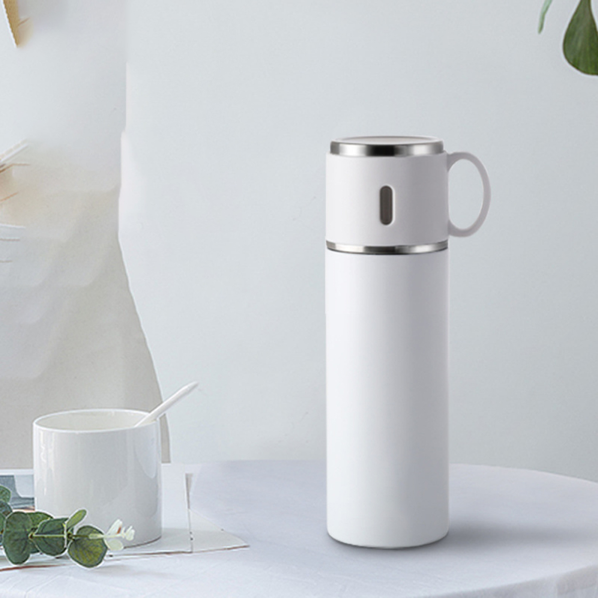 Eine weiße Thermoskanne mit einer Tasse obenauf steht auf einem weißen Tisch.