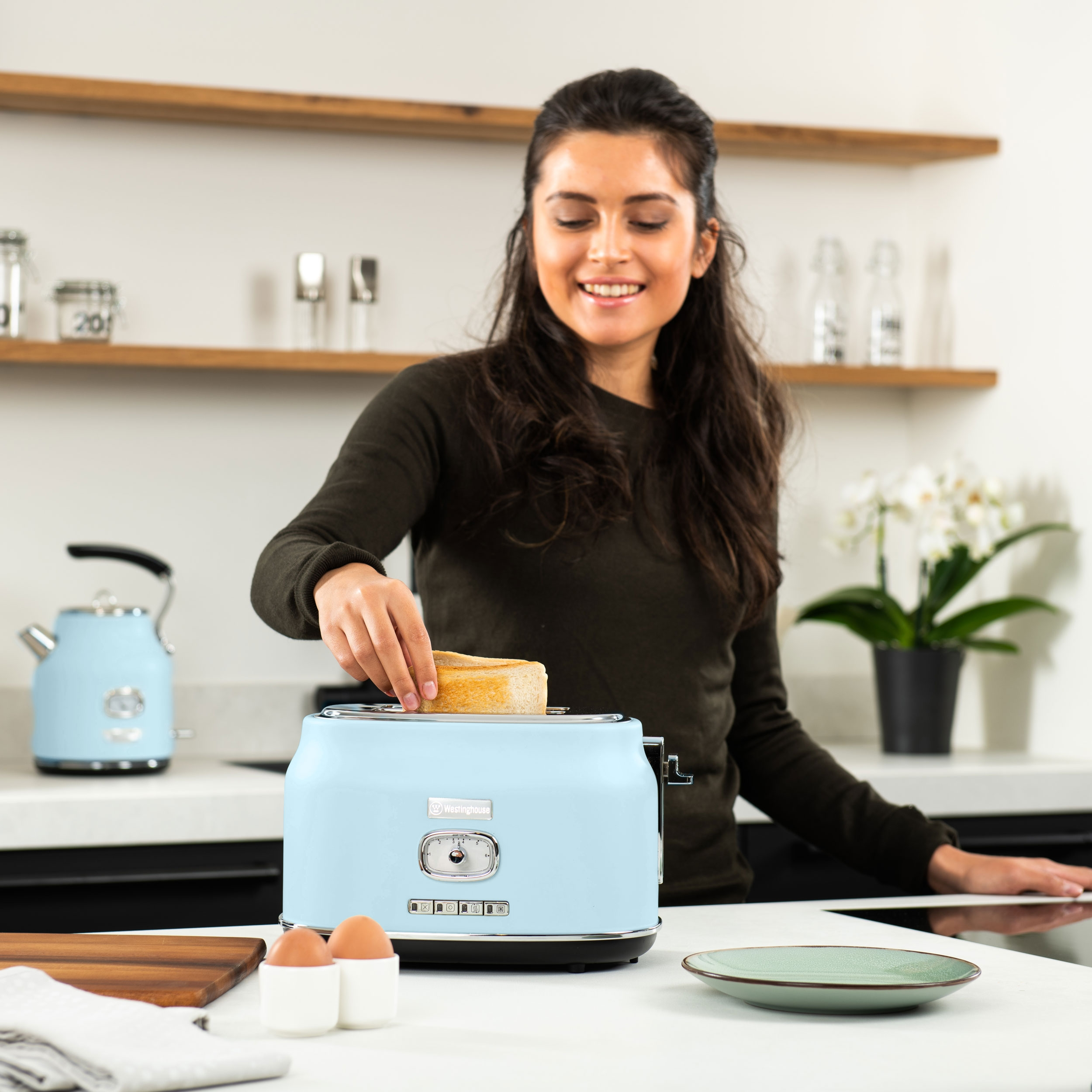 Frau legt Toast in einen blauen Toaster. Küchenszene mit Wasserkocher und Blumen.