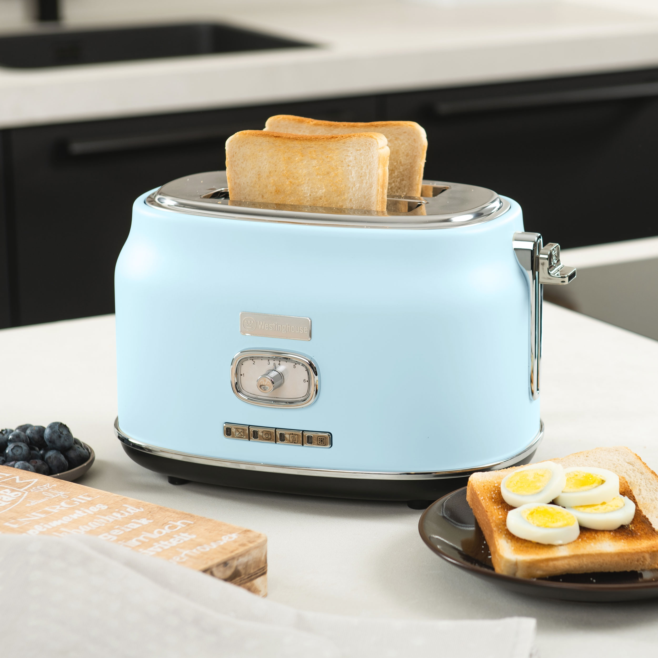Ein hellblauer Toaster mit Toast und Eiern auf einem Tisch. Es gibt Blaubeeren und eine Holzkiste.