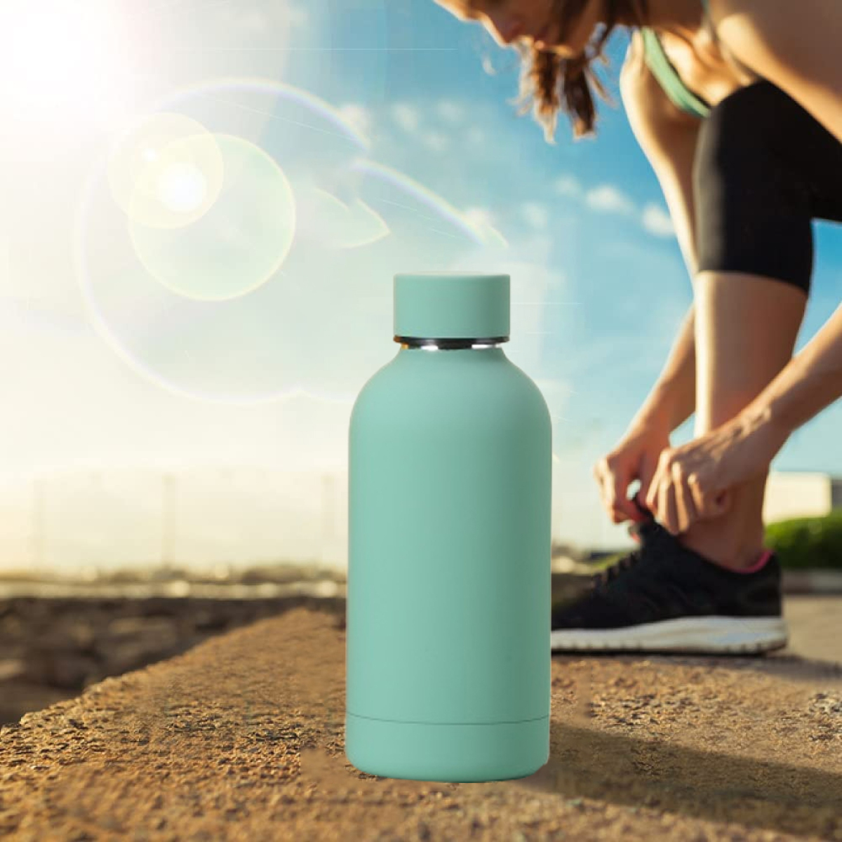 Person bindet Schnürsenkel neben mintgrüner Wasserflasche. Blauer Himmel und Sonne im Hintergrund.