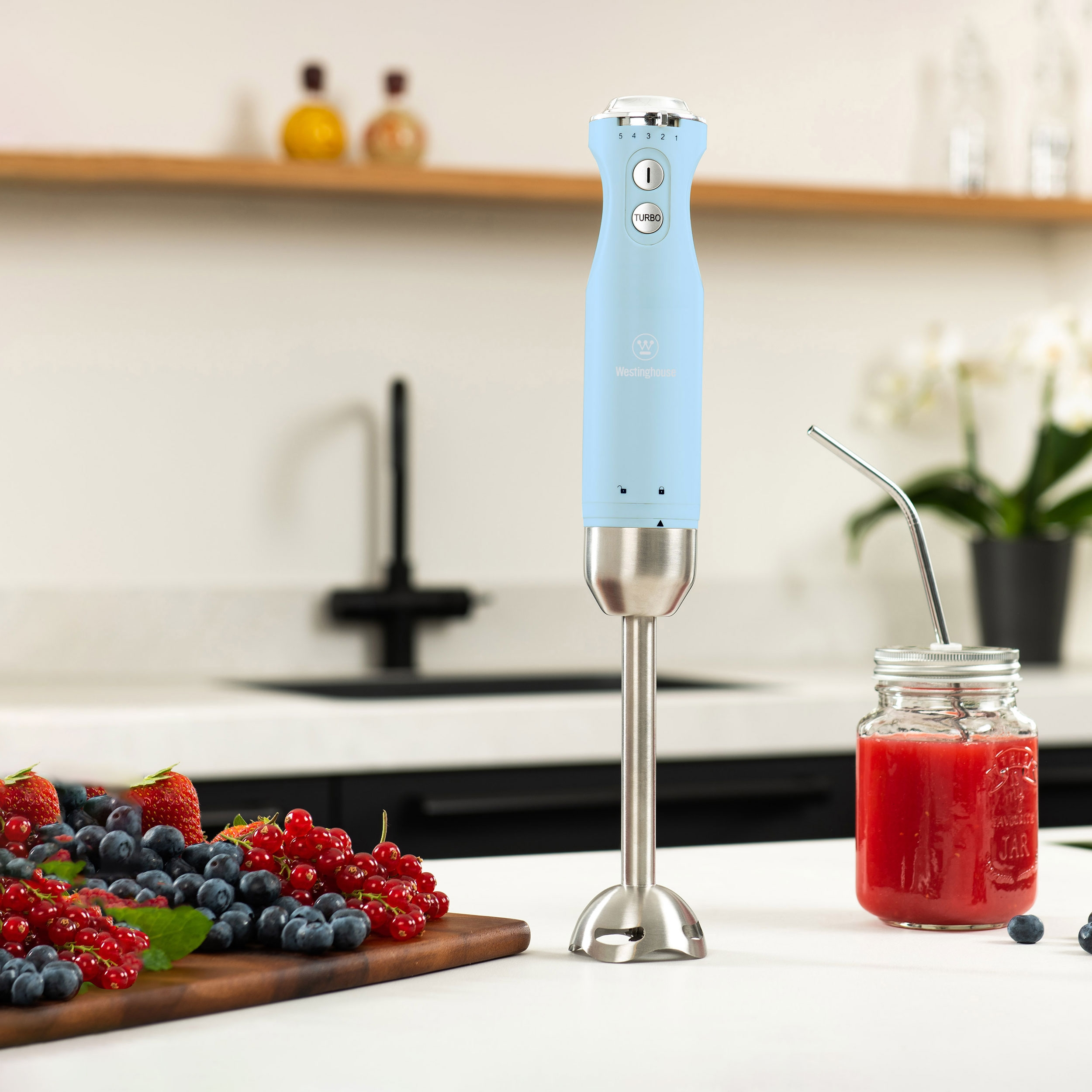 Ein hellblauer Handmixer mit einem Glas rotem Smoothie und Beeren auf einem Tisch.