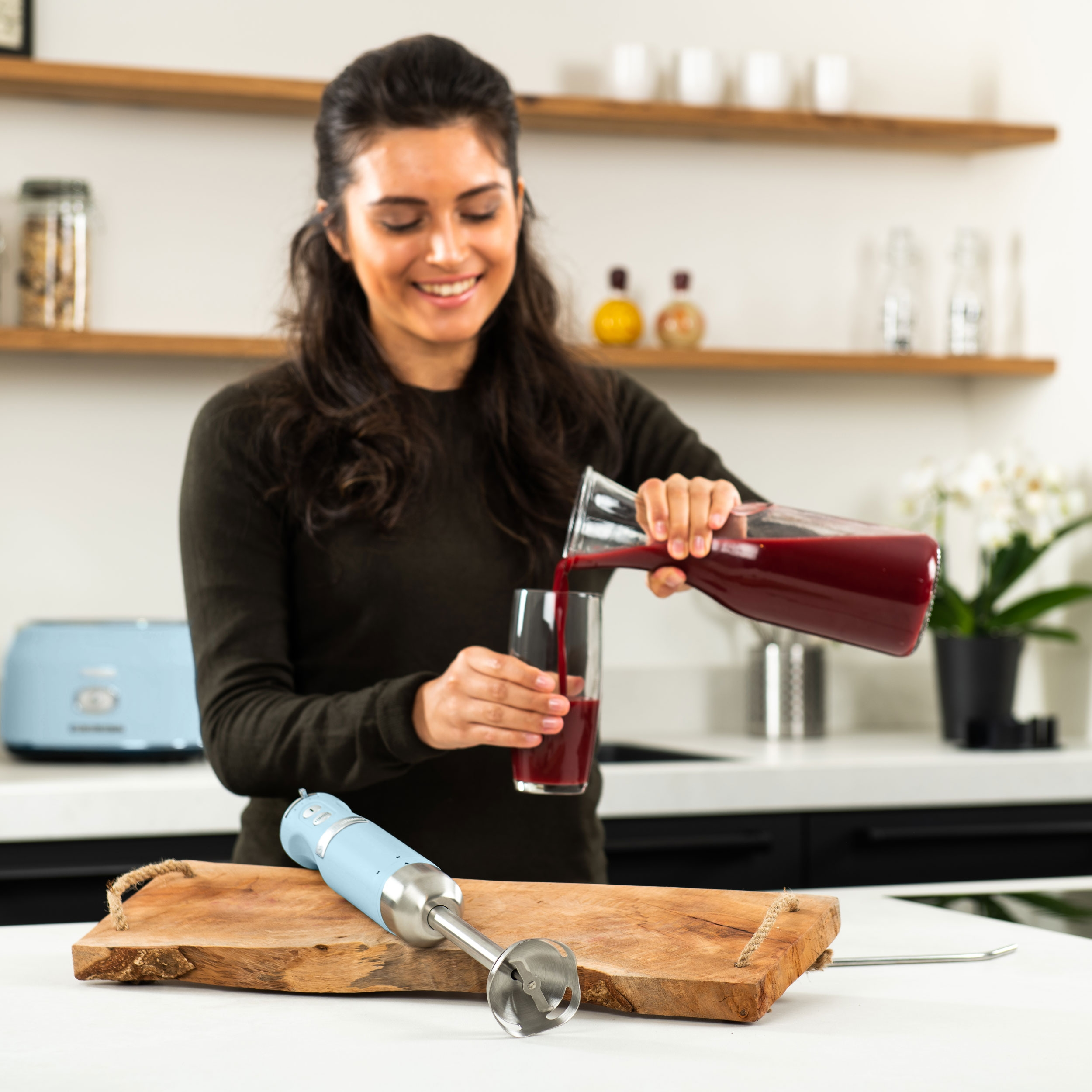 Frau gießt roten Saft in ein Glas. Ein blauer Mixer und ein Holzschneidebrett stehen auf der Theke.