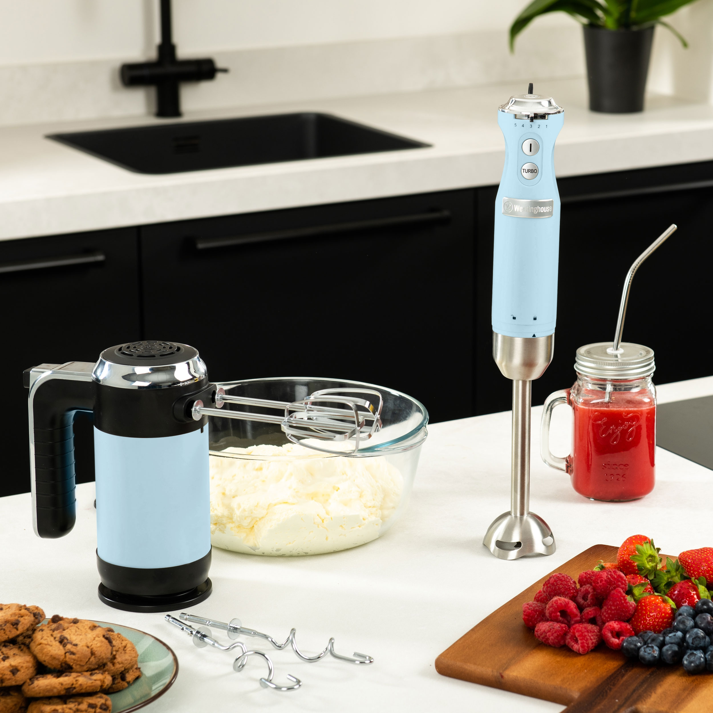 Küchenszene mit blauen Handrührern, einem Mixer, Erdbeeren und einem Glas.