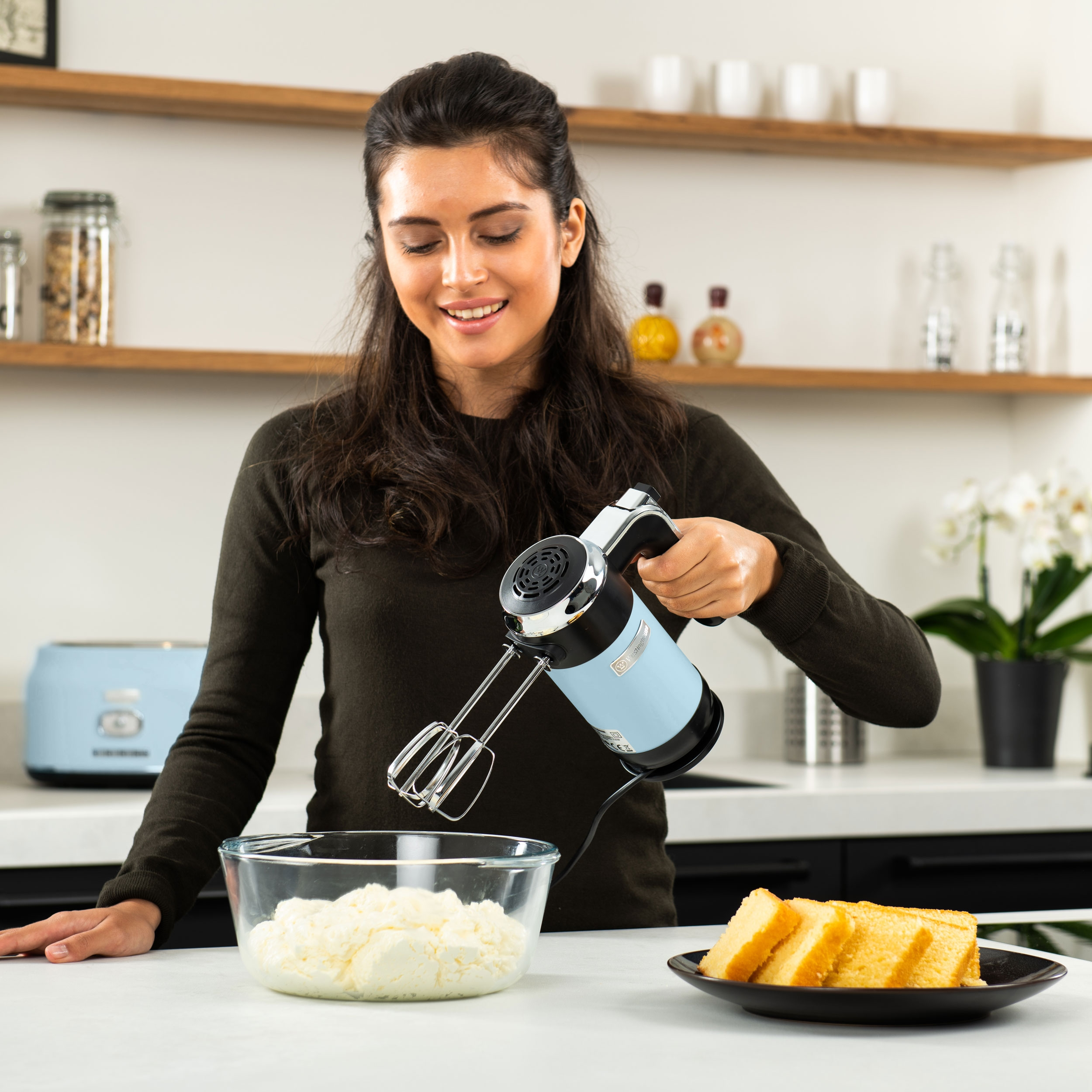 Frau verrührt Zutaten. Verwendet einen hellblauen Handmixer. Glasschüssel mit Sahne und Kuchenplatte.