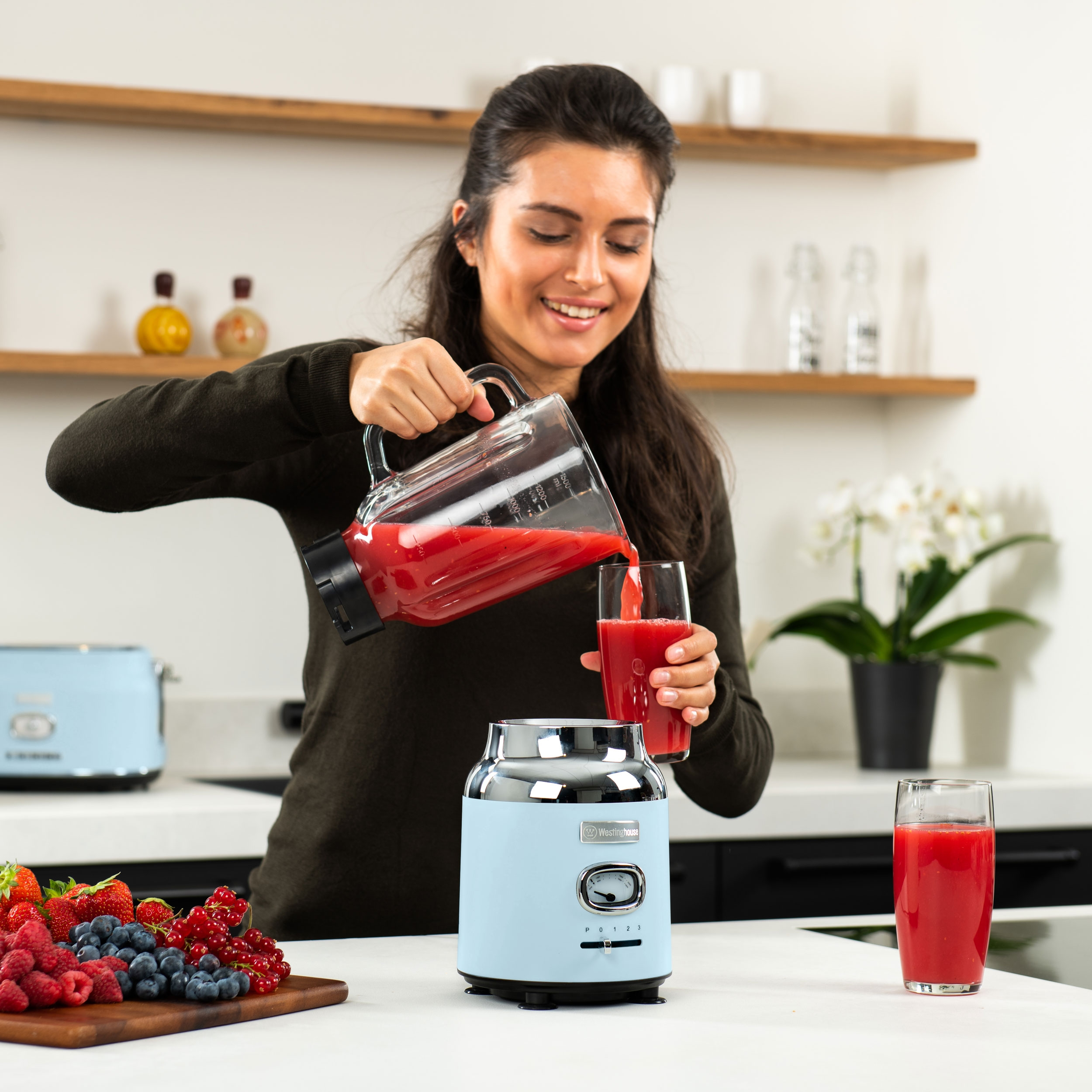 Frau gießt roten Saft in ein Glas neben einem Mixer und Früchten auf der Theke.