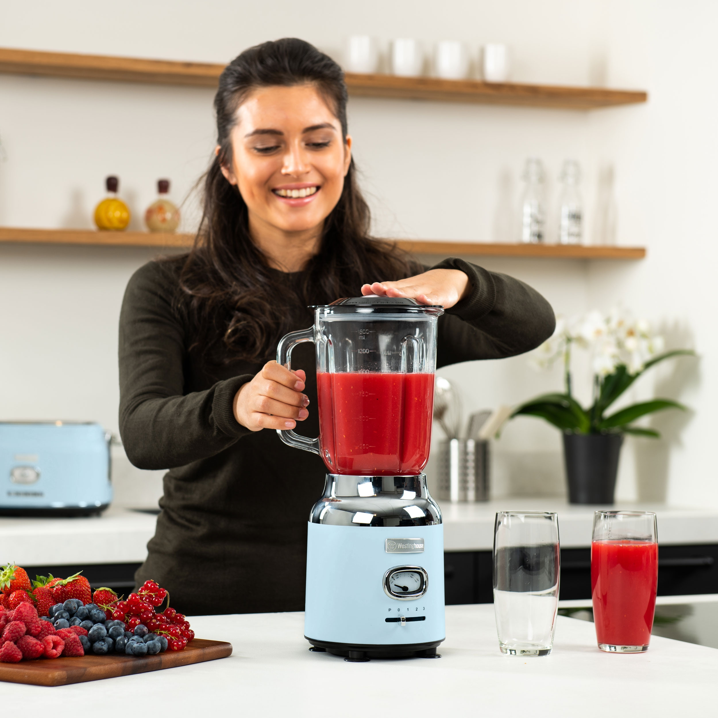 Eine Frau benutzt einen blauen Mixer mit roter Flüssigkeit, neben einem Schneidebrett mit Beeren.