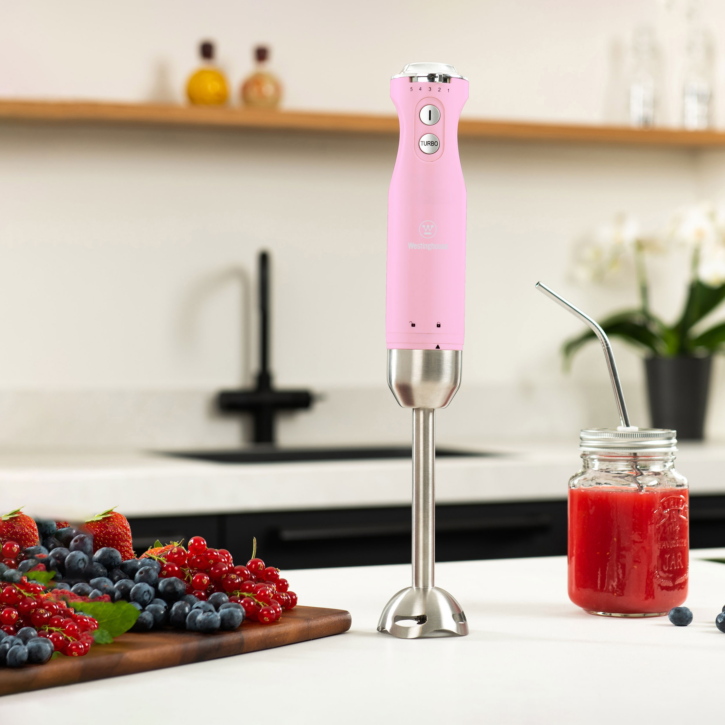 Rosa Handmixer mit Edelstahlbasis, neben rotem Smoothie und Früchten auf einem Tisch.