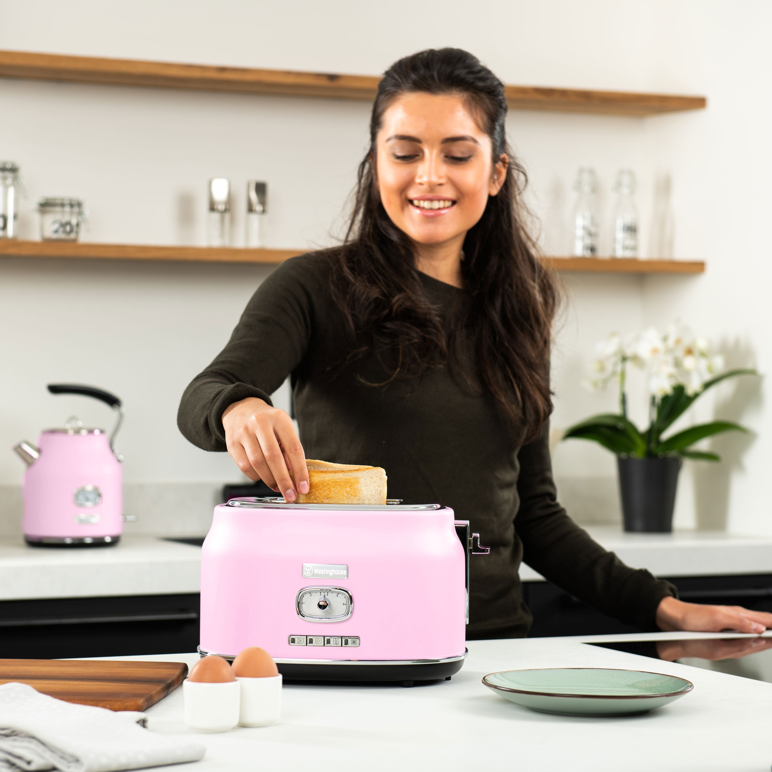 Frau legt Brot in einen rosa Toaster. Auf der Theke stehen ein Wasserkocher und Eier. Weißer Hintergrund.